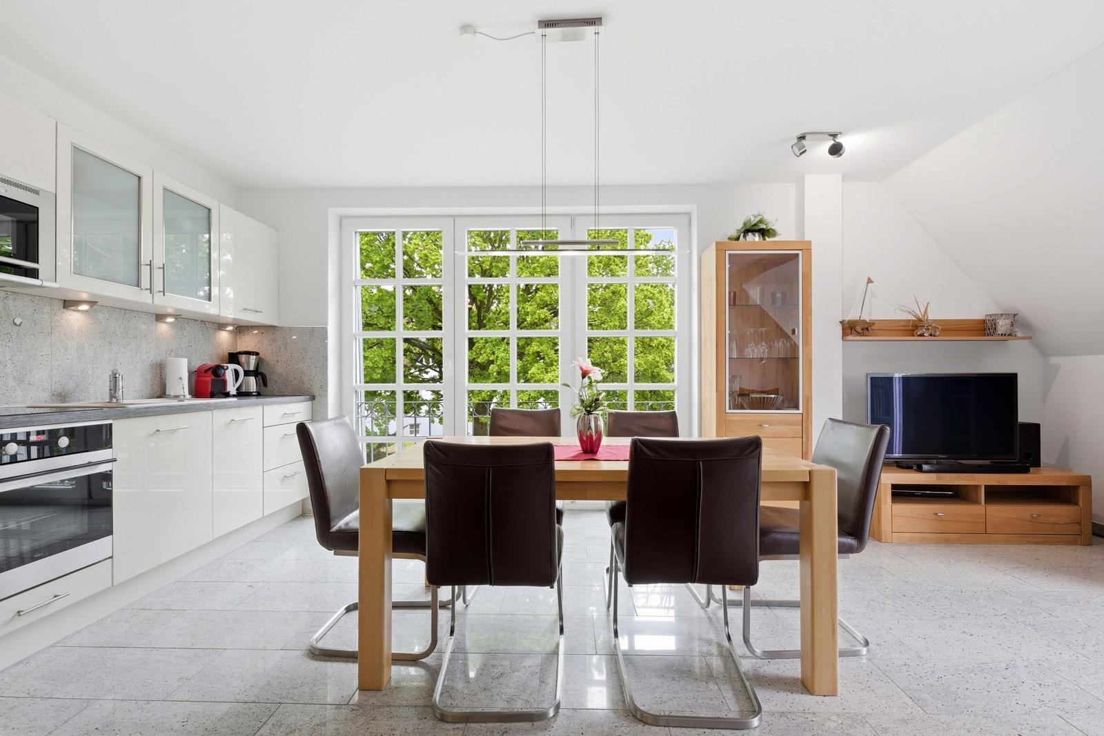 Modern kitchen with dining area and TV corner. Large windows with view of green trees.