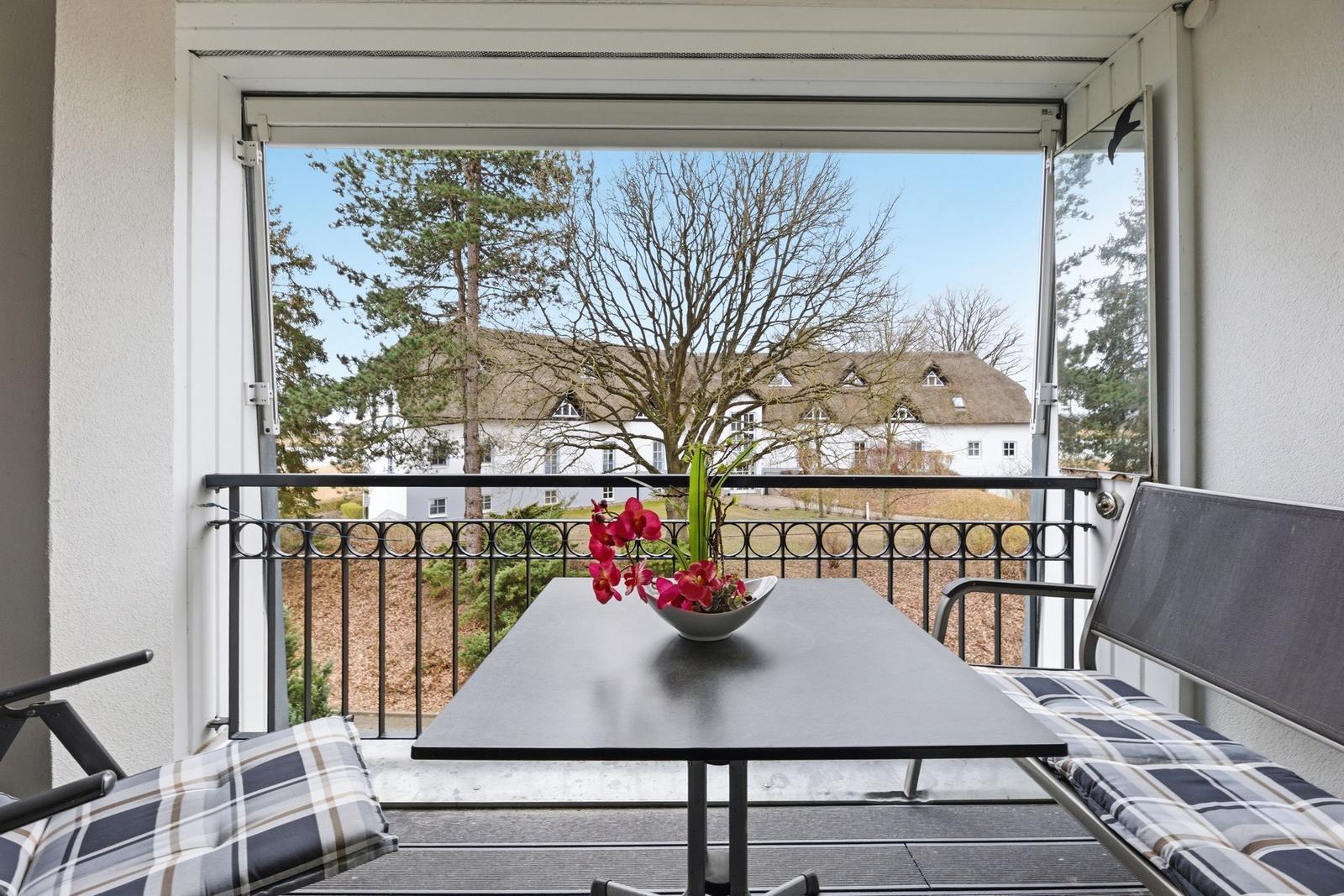 Balcony with table, chairs, and flowers. View of house and trees.