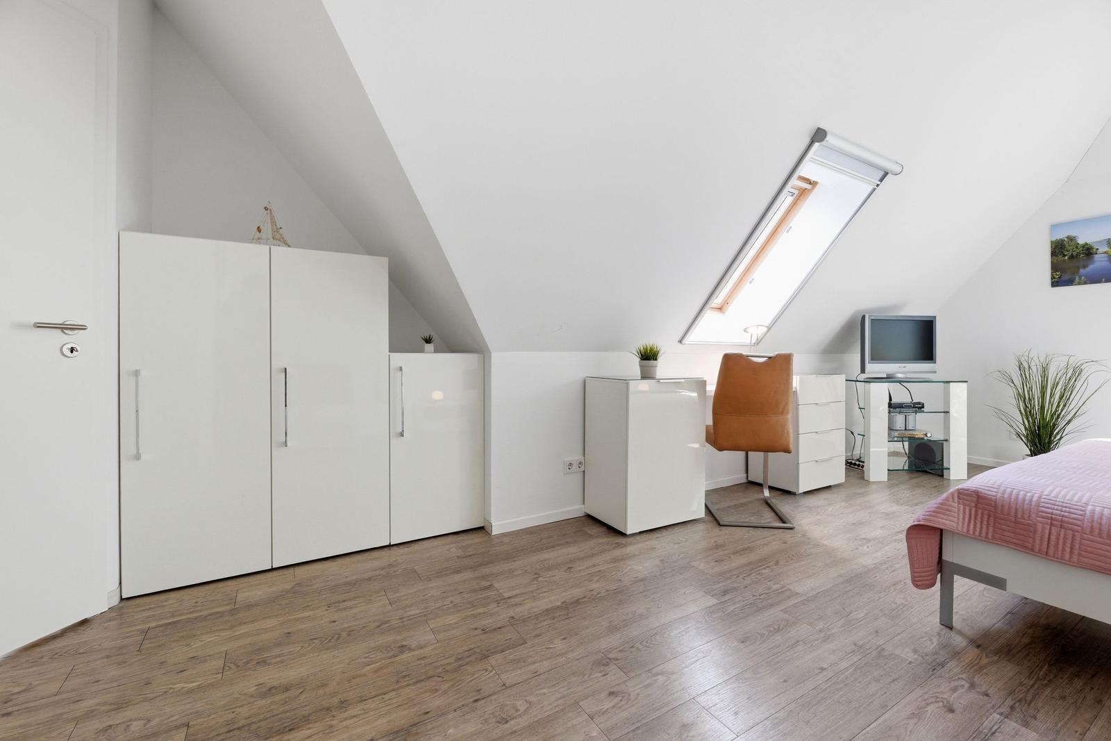 Room with sloped ceiling, white wardrobe, desk, and bed with pink bedding.