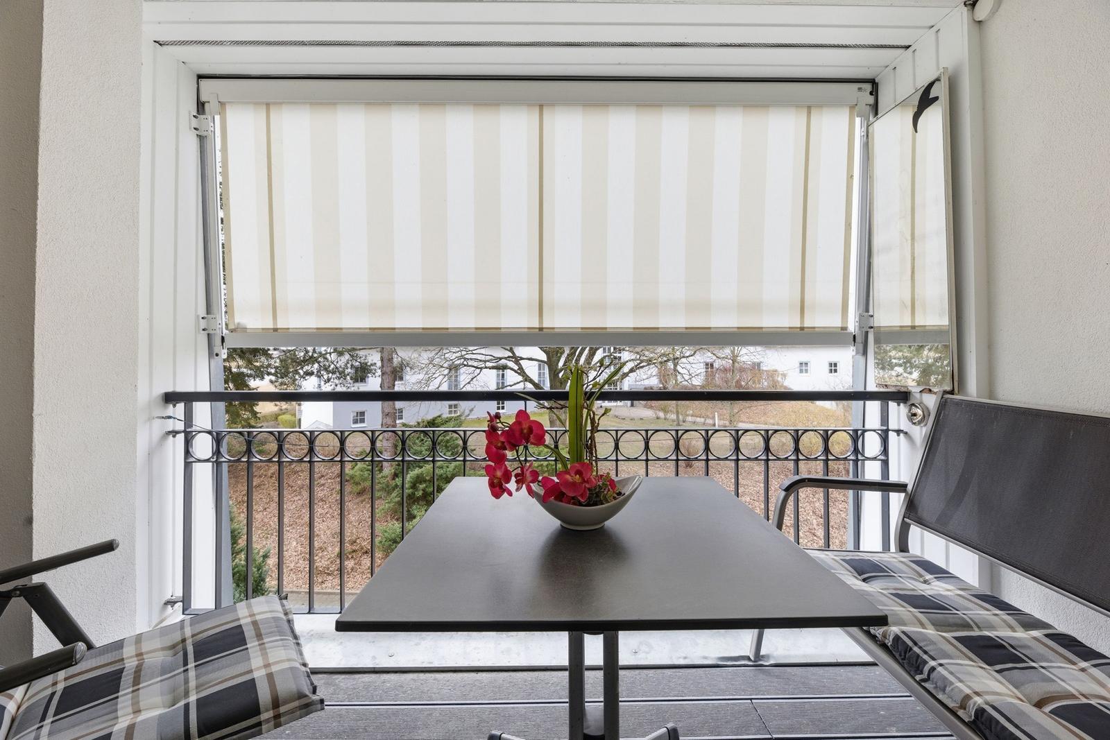 Balcony with table, chairs, and flowers. View of green areas and buildings.