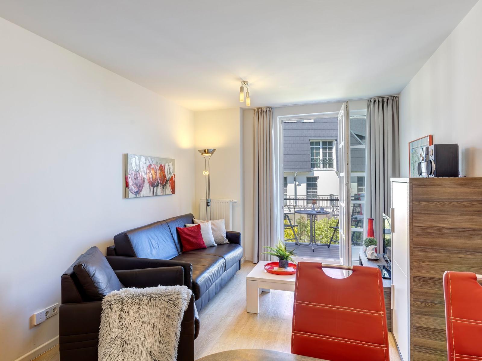 Wohnzimmer mit Sofa, Tisch und Balkonansicht. Rote Stühle und Wandbild.