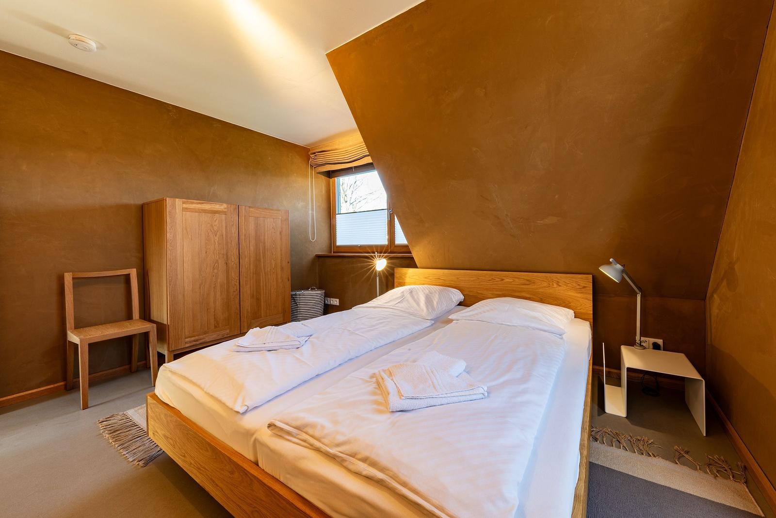 Schlafzimmer mit Bett, Nachttisch, Kleiderschrank und Stuhl. Wandfarbe braun, Fenster mit Jalousie.