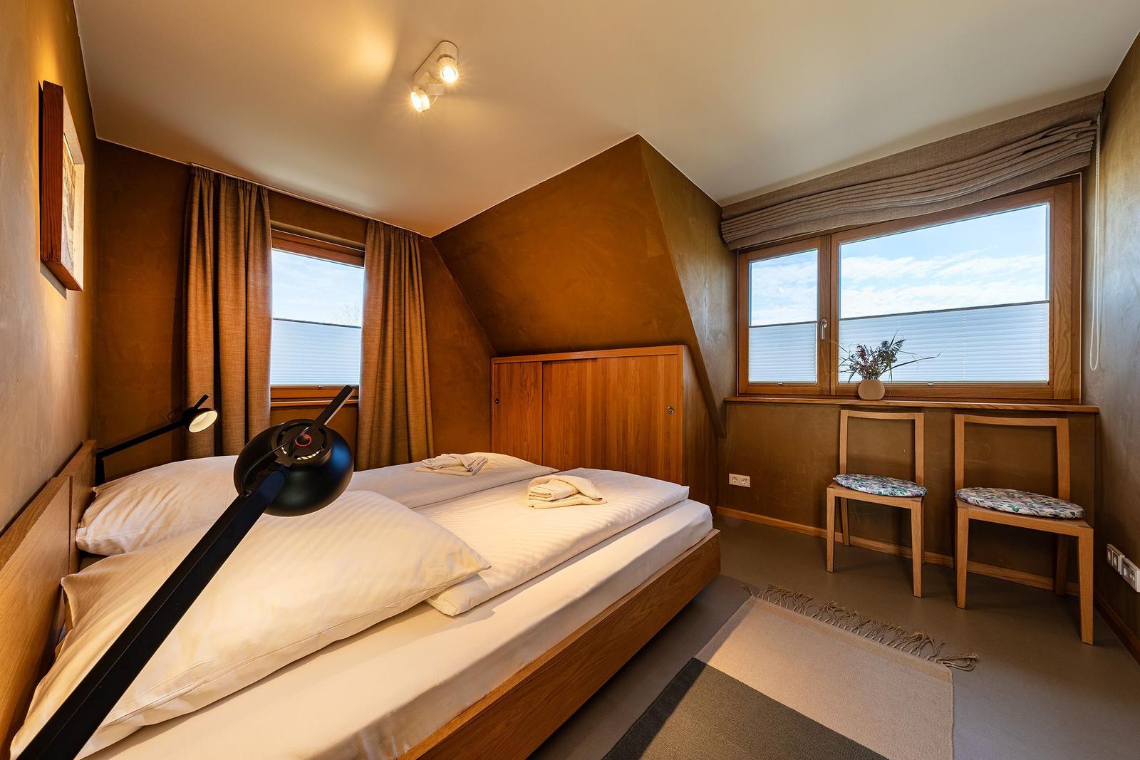 Doppelbettzimmer mit Holzmöbeln, zwei Fenstern und zwei Stühlen mit Kissen.