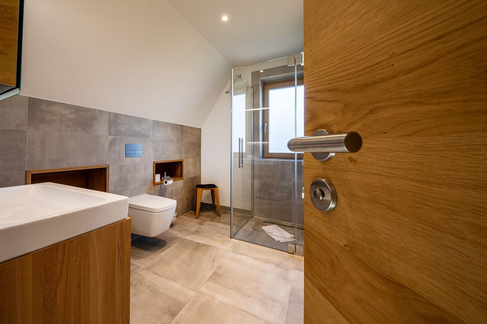 Badezimmer mit Dusche, Toilette und Waschbecken. Holztür und graue Fliesen.