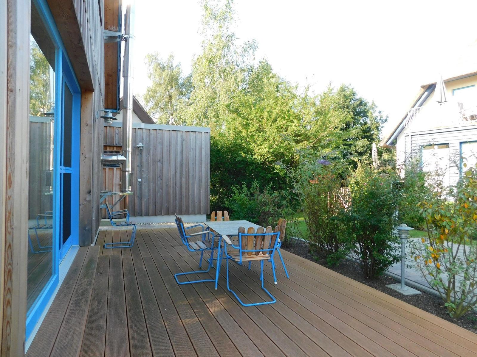 Terrasse mit Holzboden, blauen Stühlen und Tisch. Hintergrund mit Bäumen und Garten.