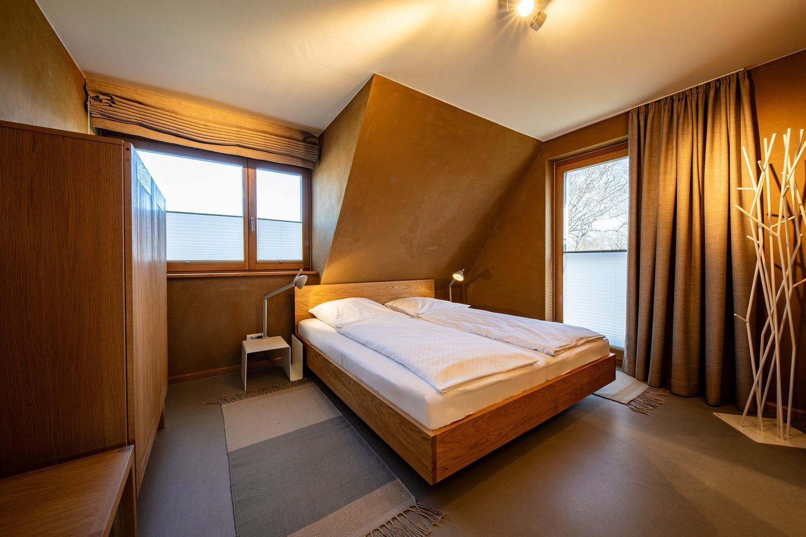 Schlafzimmer mit Doppelbett, Holzmöbeln und Fenstern mit Jalousien.