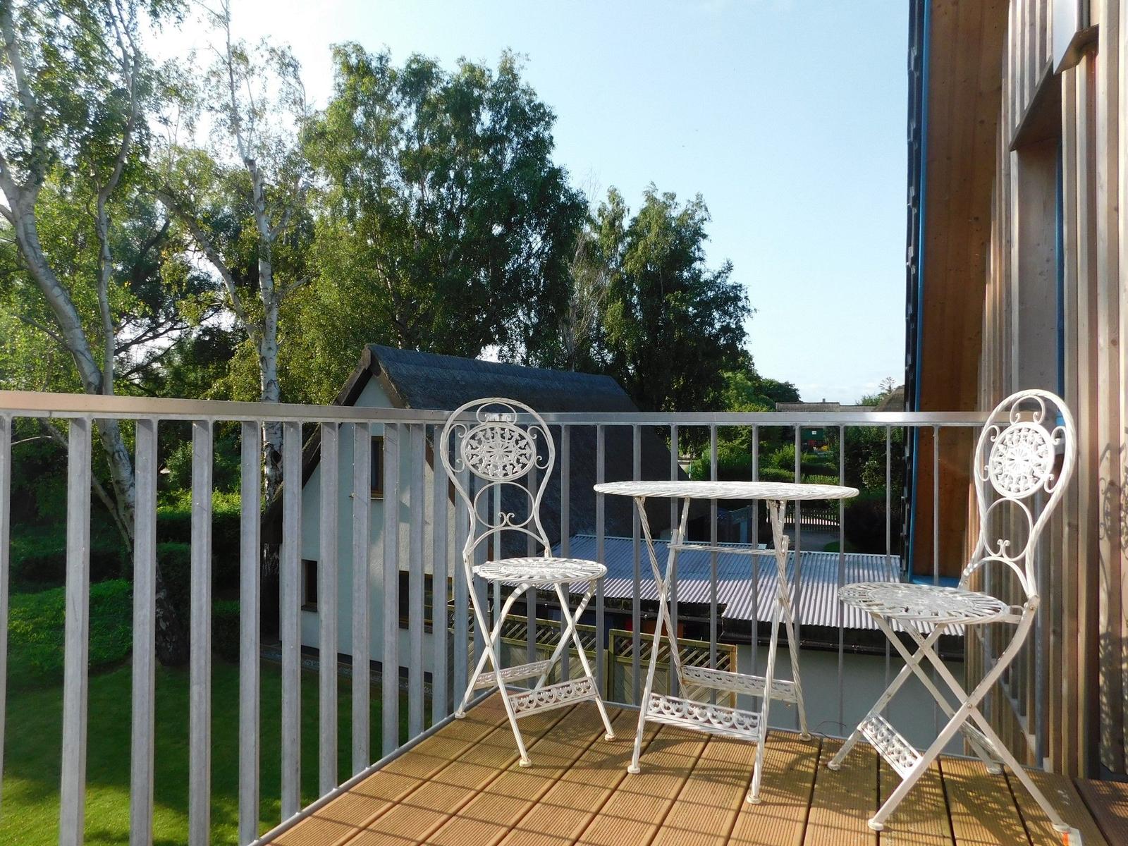 Terrasse mit weißem Tisch und zwei Stühlen. Blick auf grüne Bäume und ein Haus.