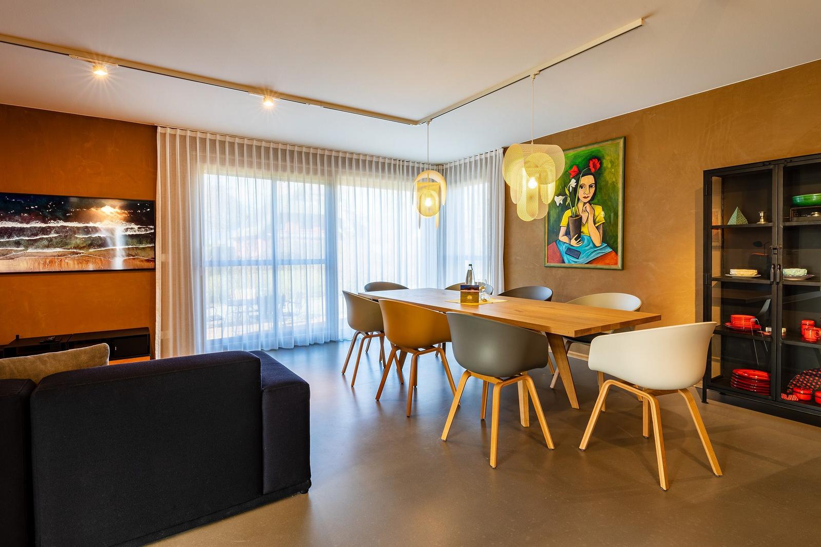 Essbereich mit Holztisch, Stühlen und Wandbildern. Fenster mit Vorhängen. Schrank mit Geschirr.