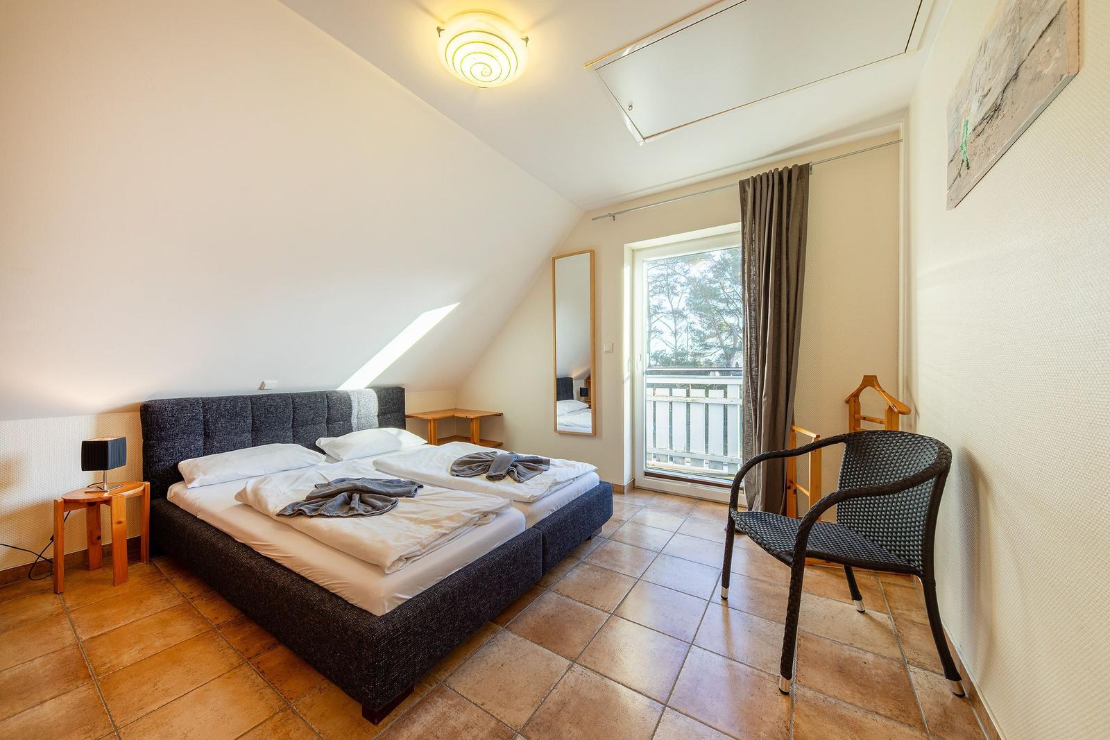 Schlafzimmer mit Doppelbett, Terrassentür und Sessel auf Fliesenboden.