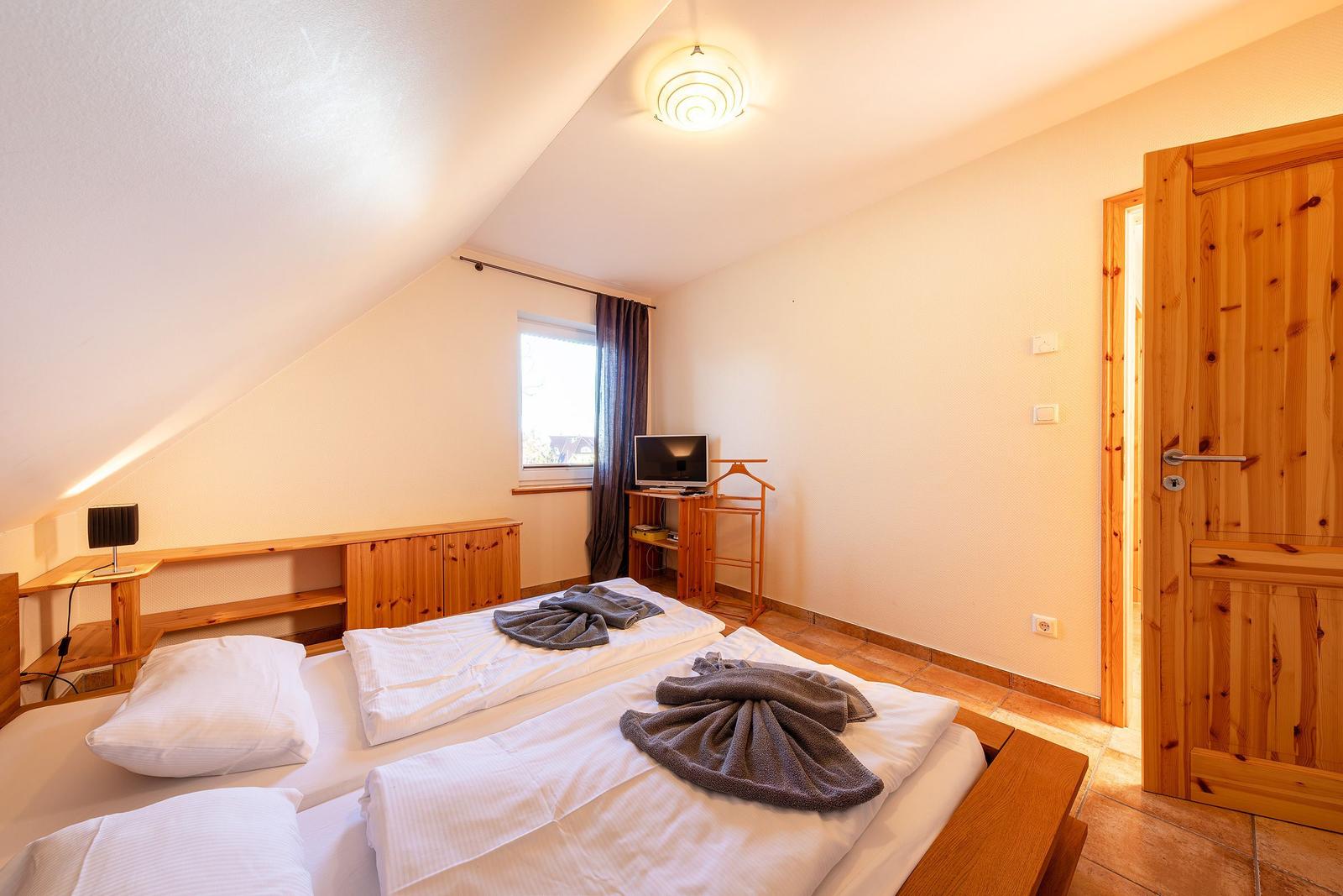 Doppelbettzimmer mit Holzmöbeln, Fenster und TV. Tür führt in Bad.