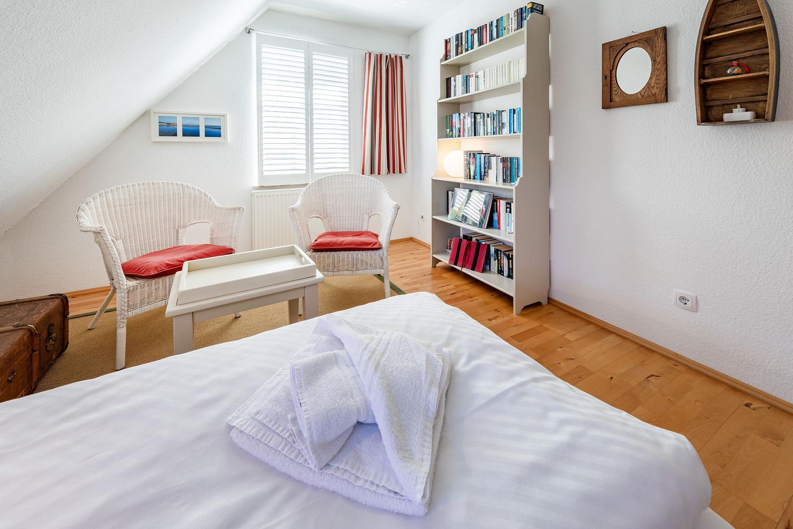 Zimmer mit Bett, zwei Stühlen, Bücherregal und Fenster mit Jalousie.