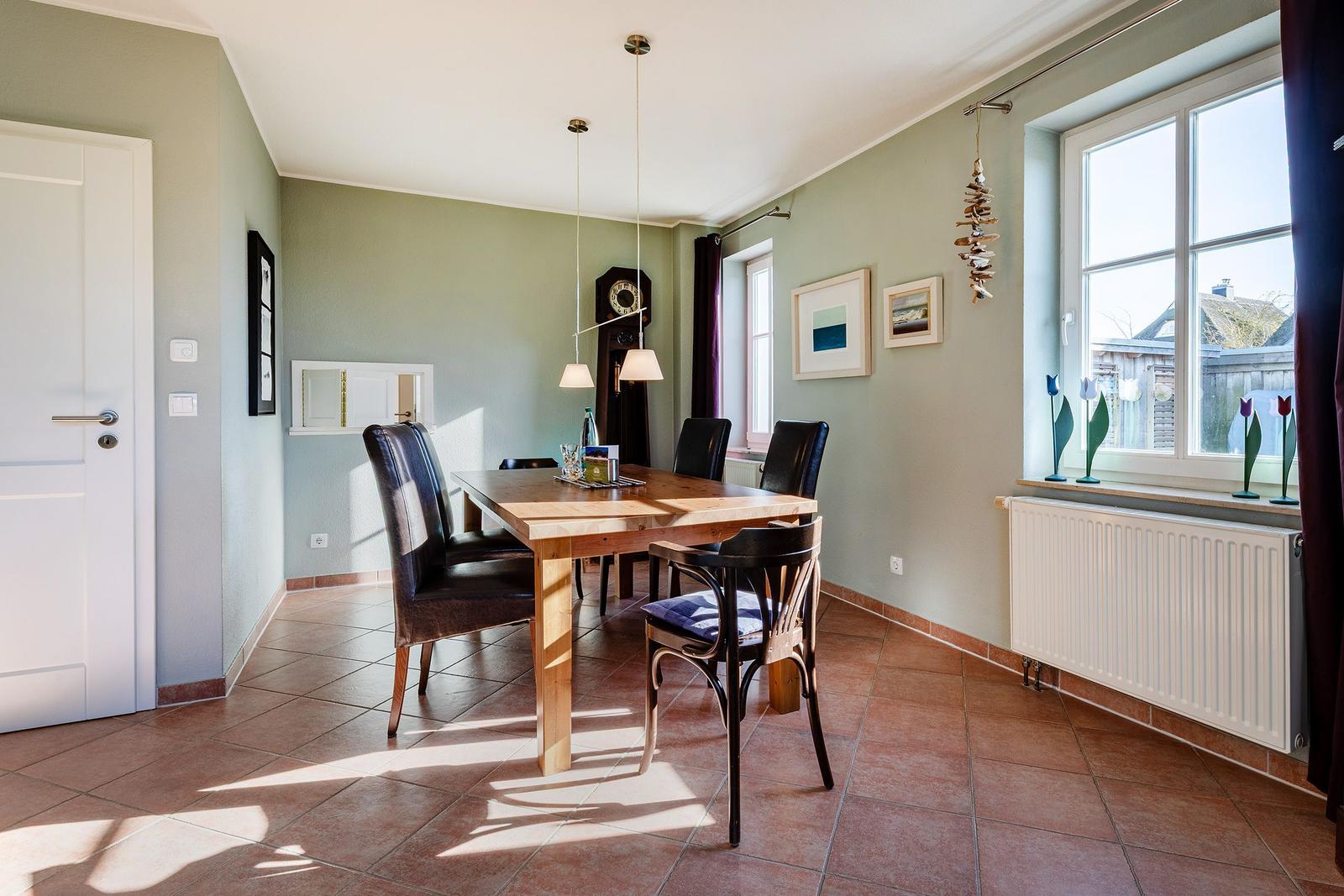 Esszimmer mit Holztisch, Stühlen und großen Fenstern. Wanduhr und Bilder an den Wänden.