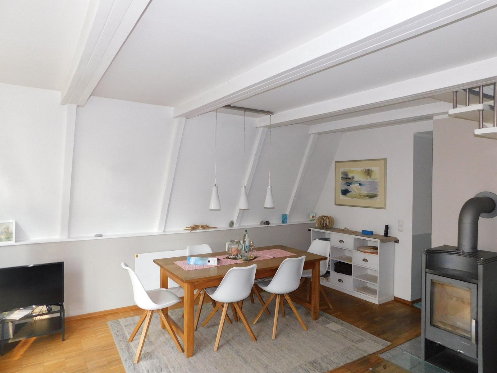 Esszimmer mit Holztisch, weißen Stühlen und Kaminofen. Deckenbalken und Wandbild sichtbar.