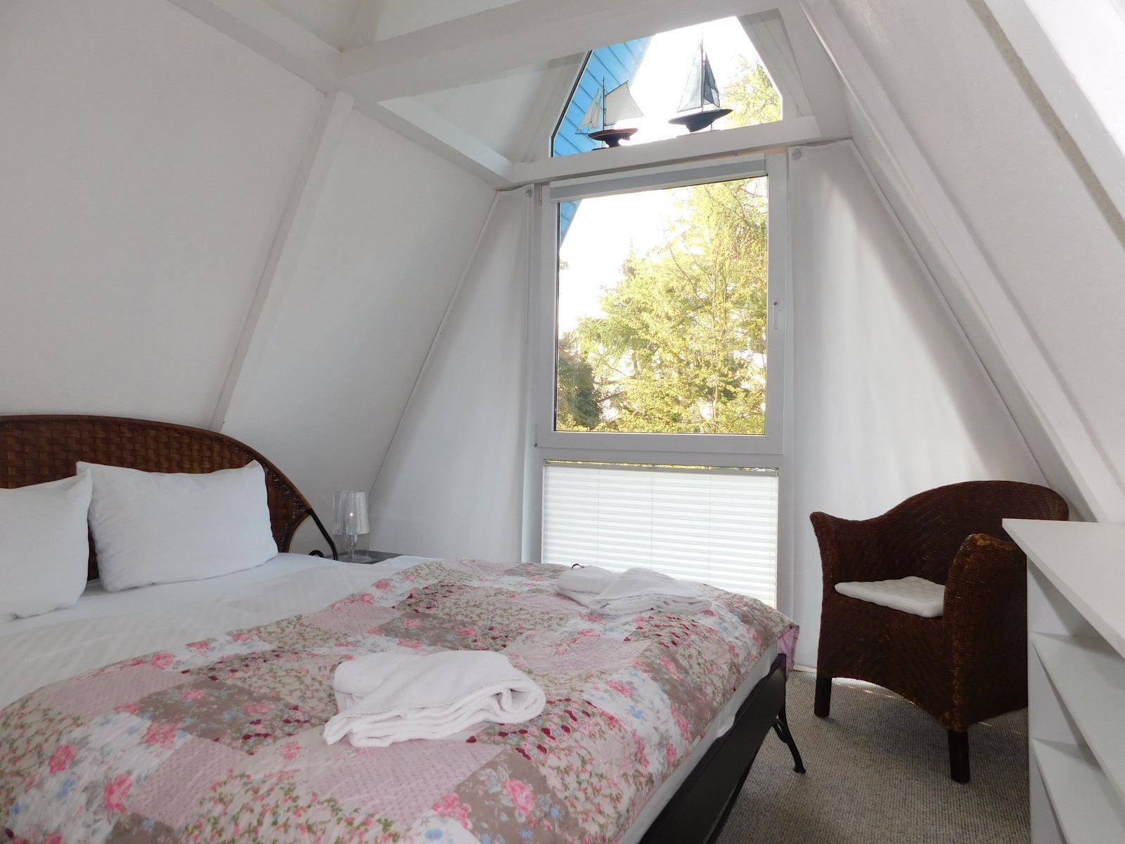Schlafzimmer mit Dachschrägen, Bett, Stuhl und Fenster mit Blick auf Bäume.
