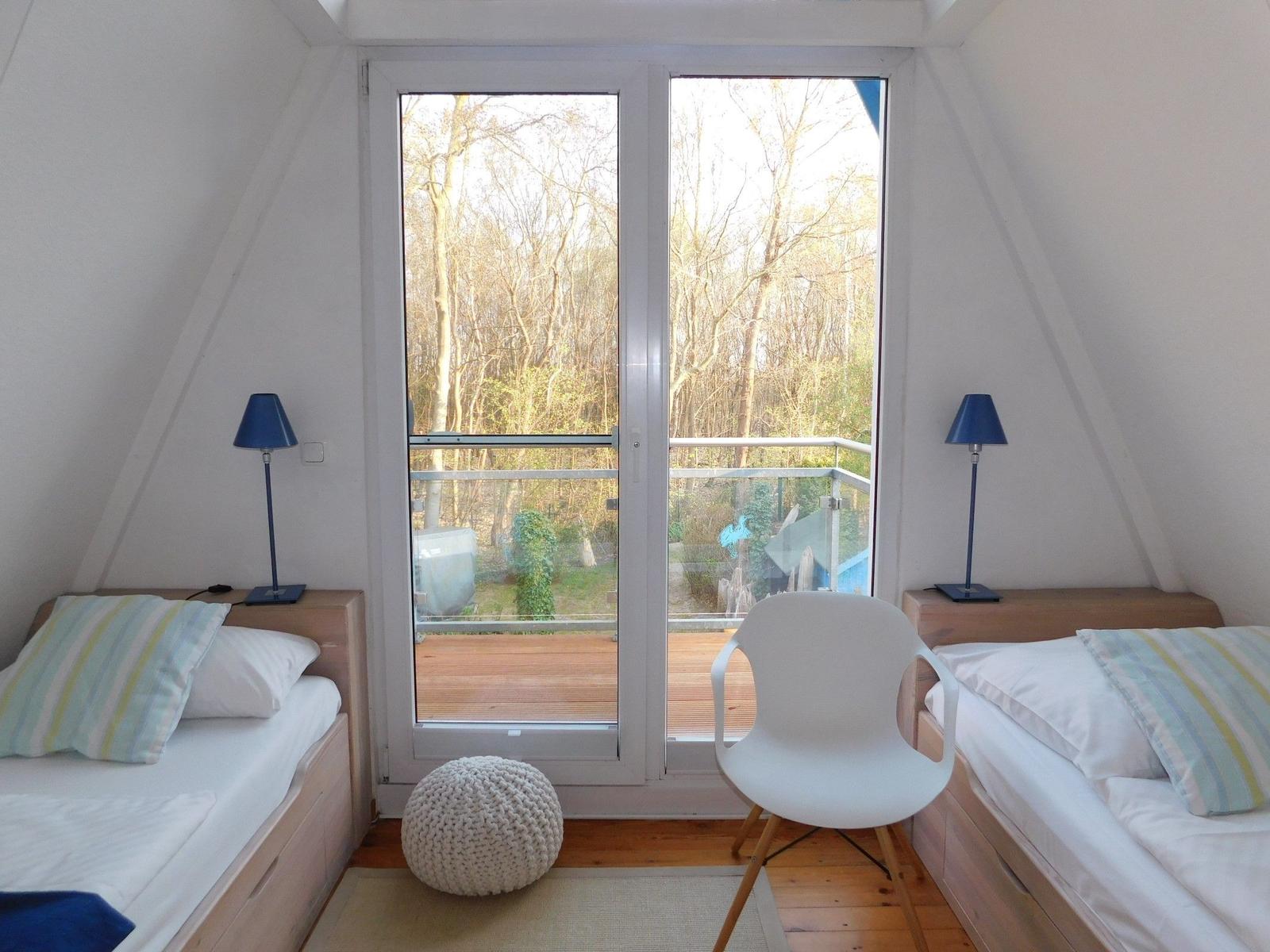 Zwei Betten mit Streifenkissen, weiße Stuhle und blau leuchtende Lampen vor großem Fenster mit Blick auf Wald.