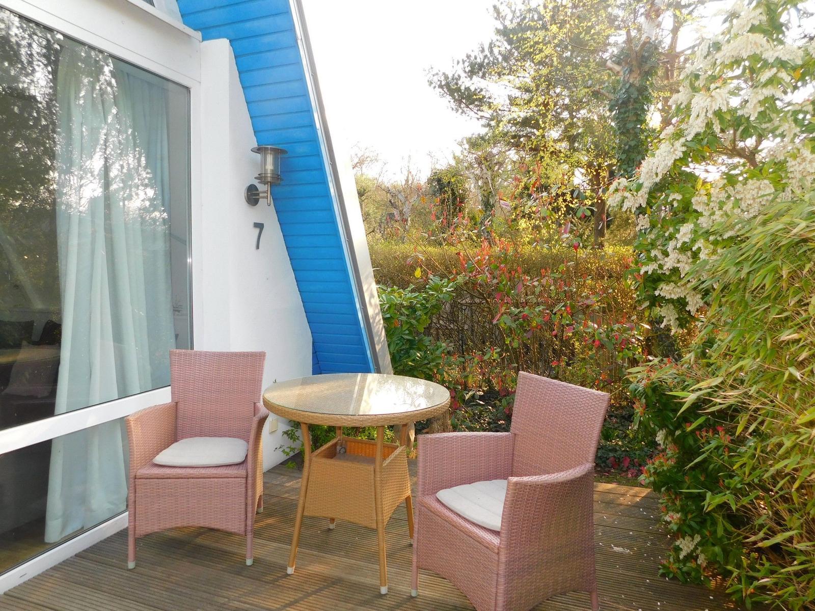 Terrasse mit Tisch und zwei Stühlen, umgeben von Pflanzen und einem blauen Gebäude.