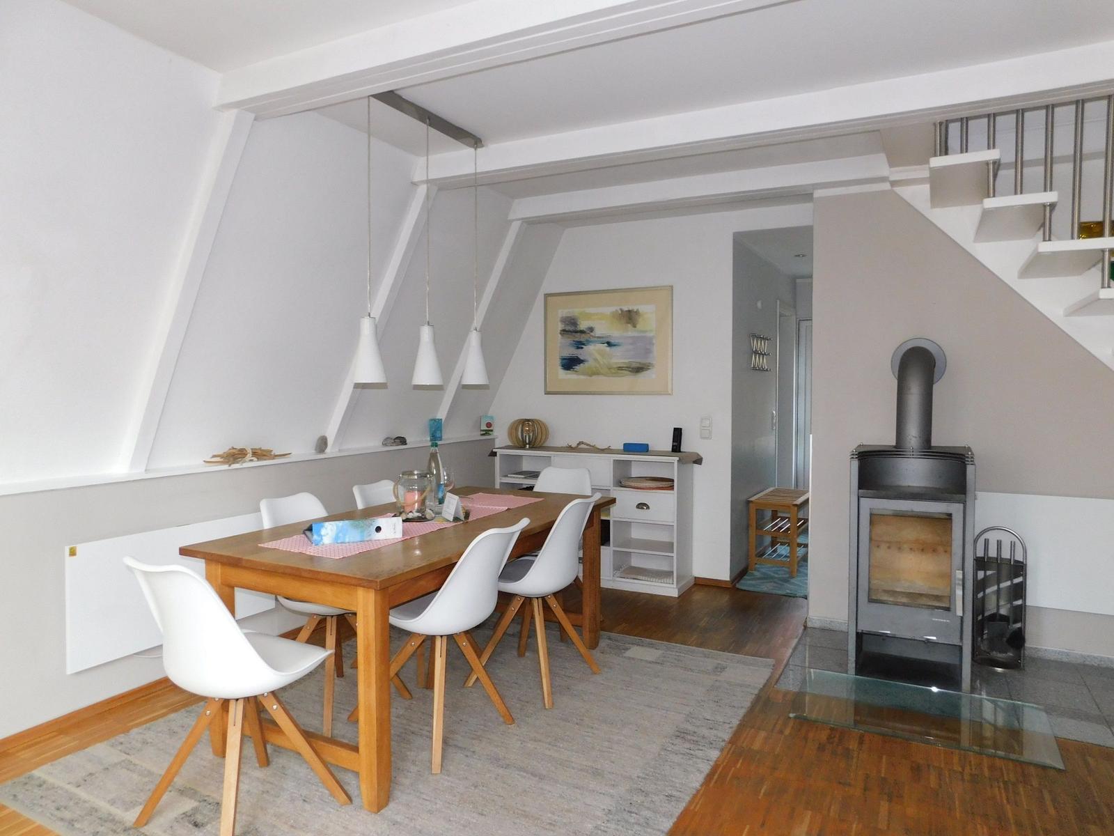 Essbereich mit Holztisch, weißen Stühlen und Kaminofen. Dachschräge und Treppe sind sichtbar.
