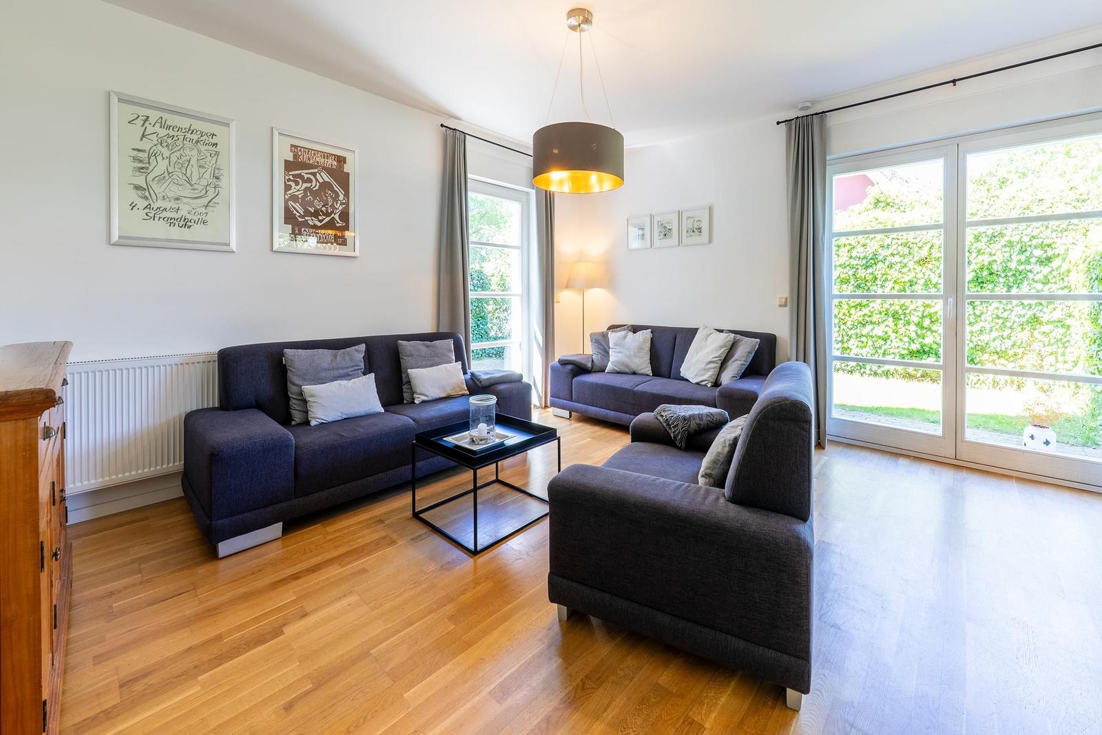 Wohnzimmer mit zwei Sofas, Tisch und großen Fenstern. Holzboden und Wandbilder.