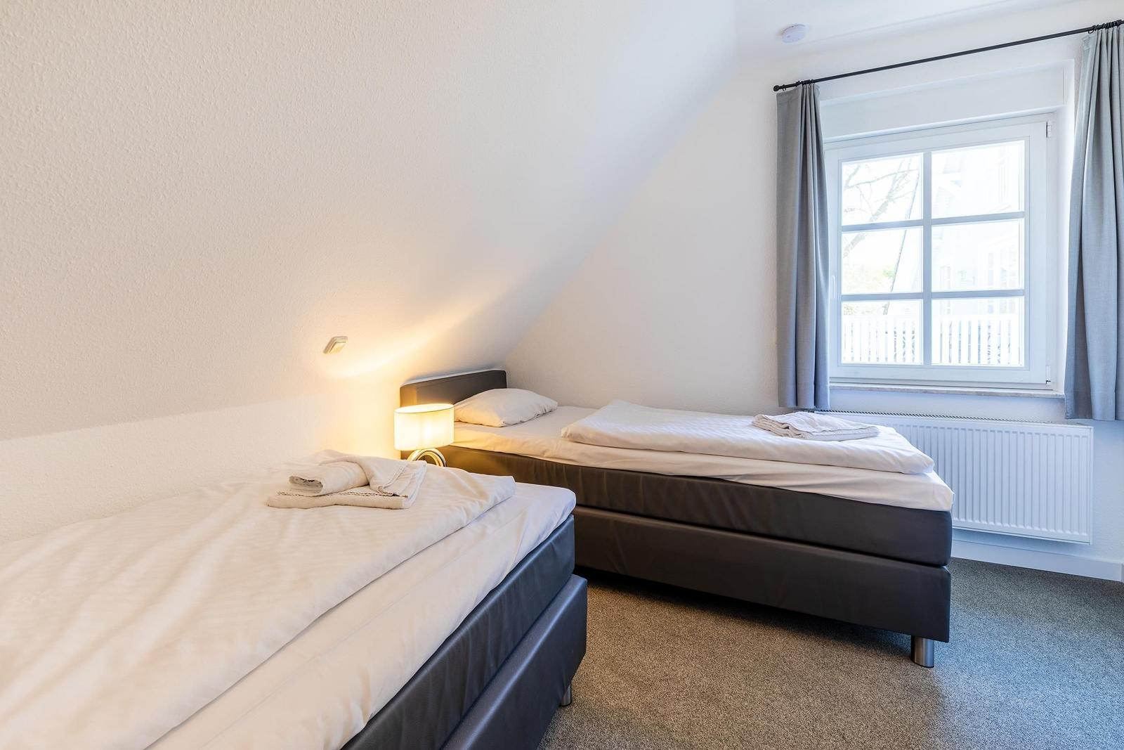 Zwei Einzelbetten mit weißen Bettwäschen und Handtüchern. Fenster mit grauen Vorhängen.