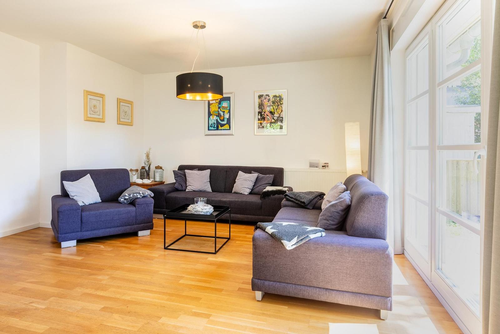 Wohnzimmer mit Sofa, Sessel und Holzboden. Große Fenster und Deckenleuchte.