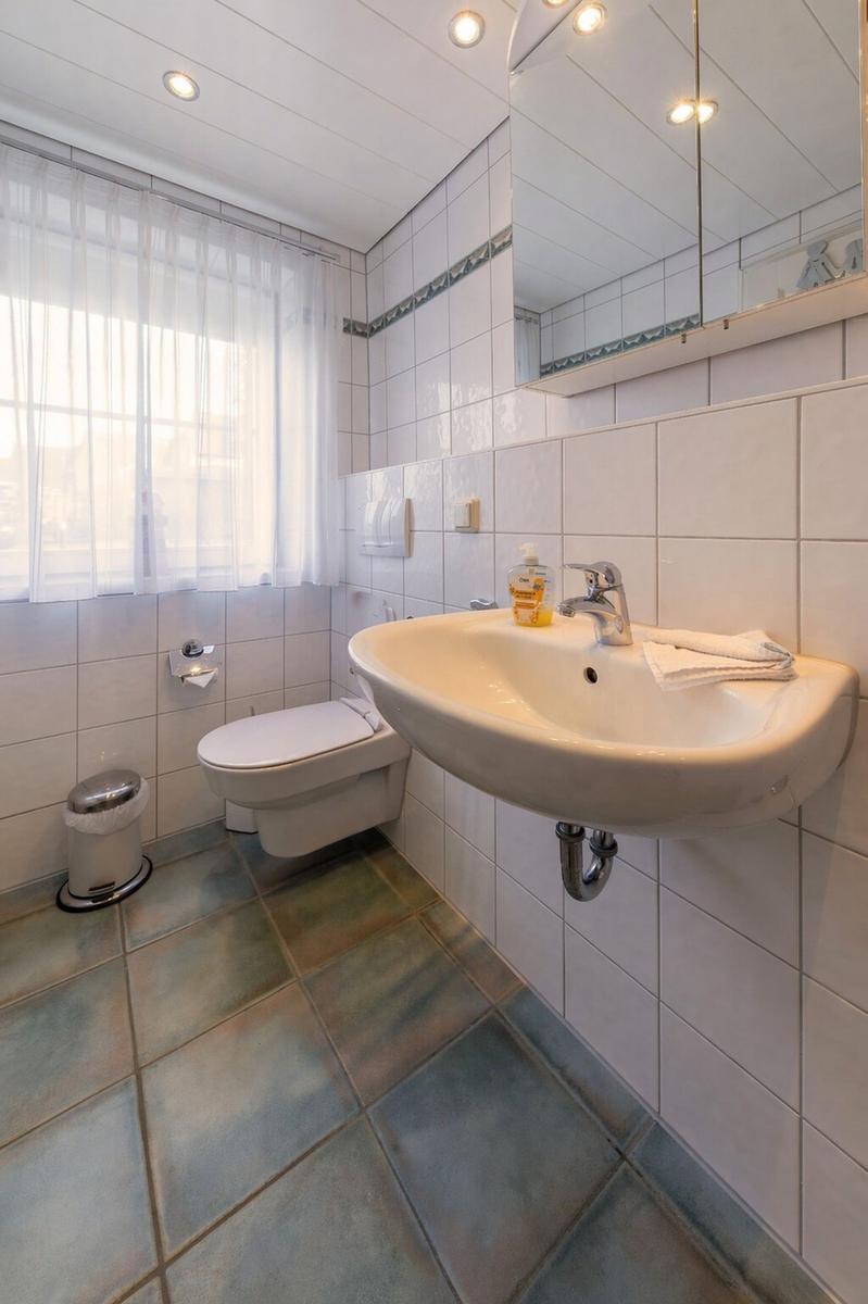 Badezimmer mit Waschbecken, Spiegel, WC und Fenster mit Vorhang.