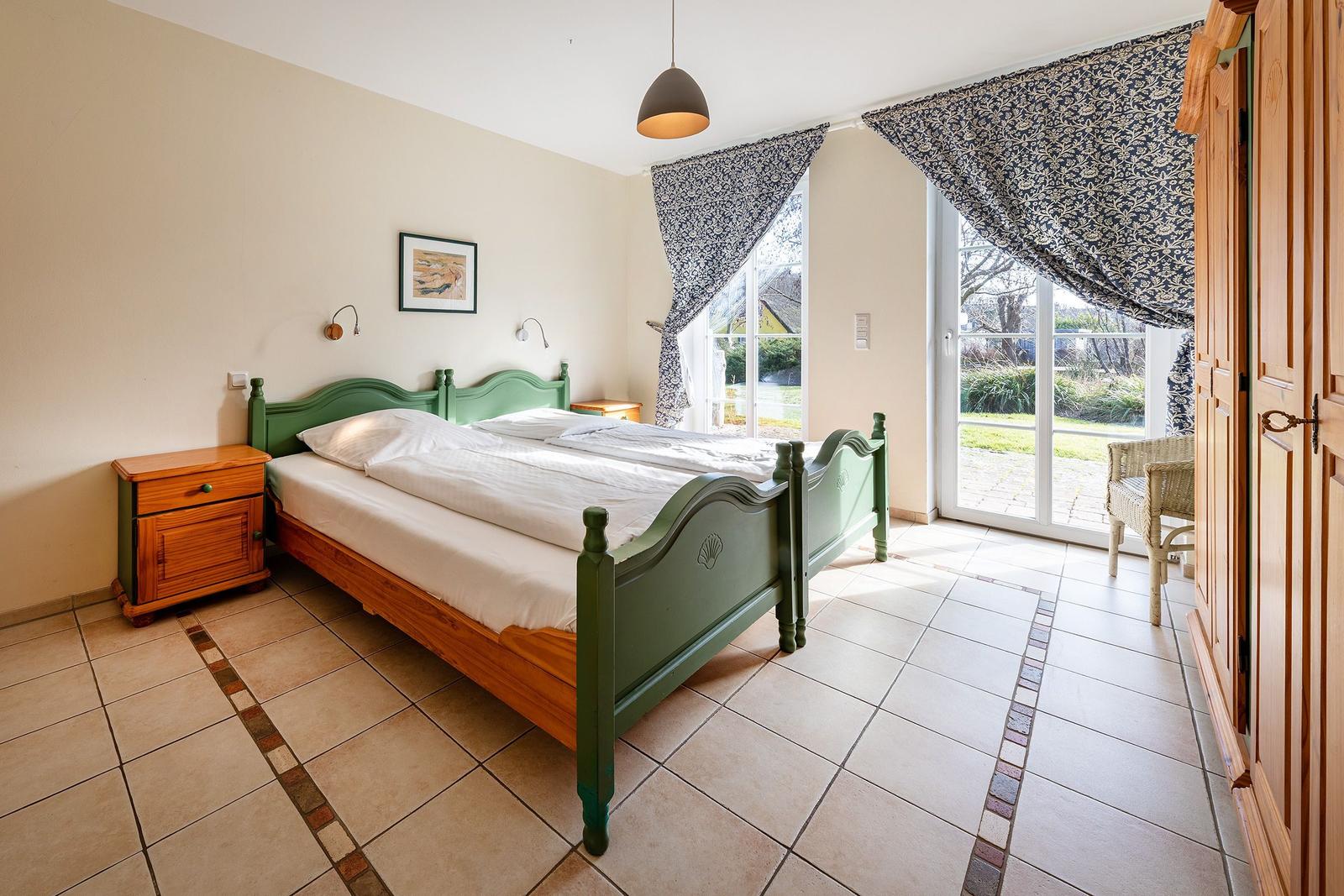 Schlafzimmer mit Bett, Nachttisch und Terrassentür. Holzmöbel und Fliesenboden.