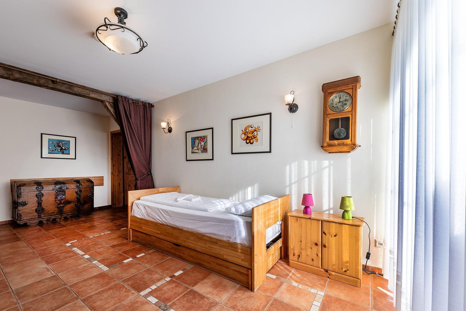 Schlafzimmer mit Holzbett, Wanduhr und Holzschrank. Terrakottafußboden und Bilder an der Wand.
