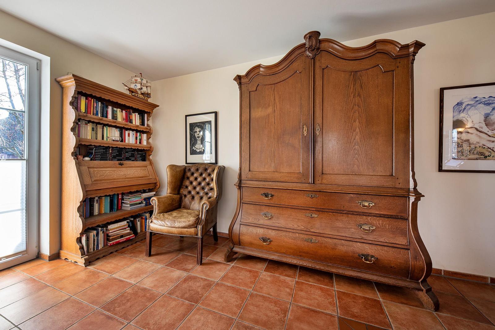 Raum mit Holzschrank, Bücherregal und Ledersessel auf Terrakottaboden.