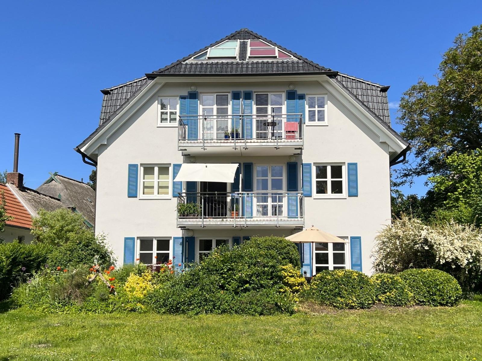 Ein zweistöckiges Haus mit Balkonen und blauen Fensterläden. Umgeben von Grün und Blumen.