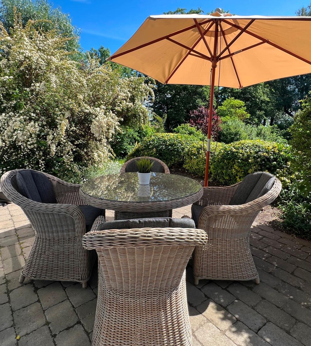 Außenbereich mit Rattan-Sitzgruppe, Glas-Tisch und Sonnenschirm unter blauem Himmel.