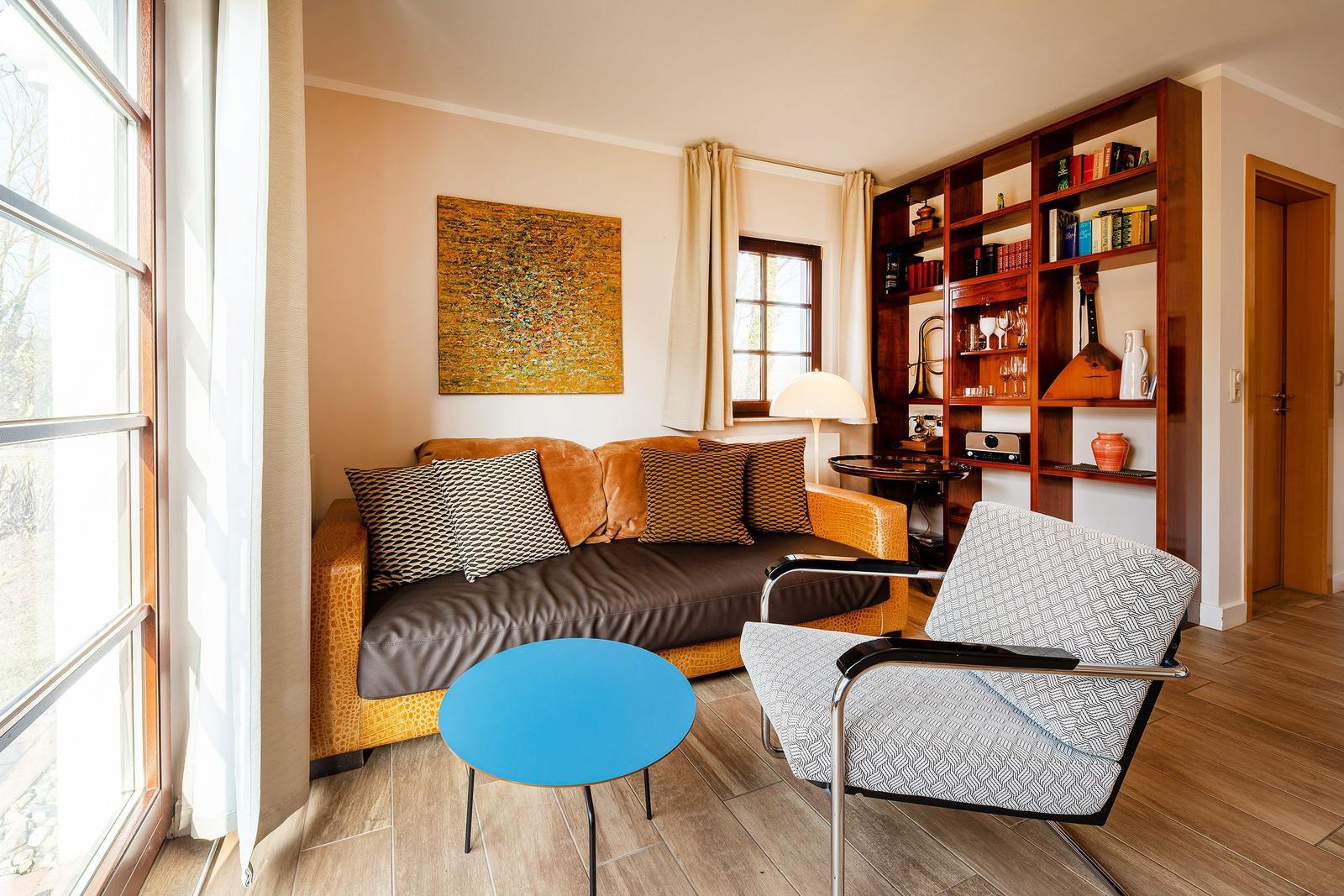 Wohnzimmer mit Sofa, Sessel, Tisch und Bücherregal. Fenster mit Vorhängen.