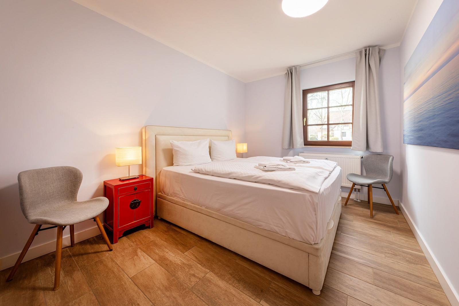 Schlafzimmer mit Bett, zwei Stühlen und rotem Nachttisch. Fenster mit Vorhängen und Wandbild.