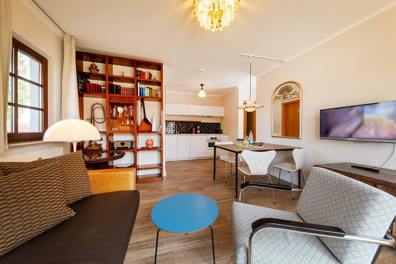 Wohnbereich mit Essplatz, Küche und Bücherregal. Deckenleuchte und Fernseher sichtbar.