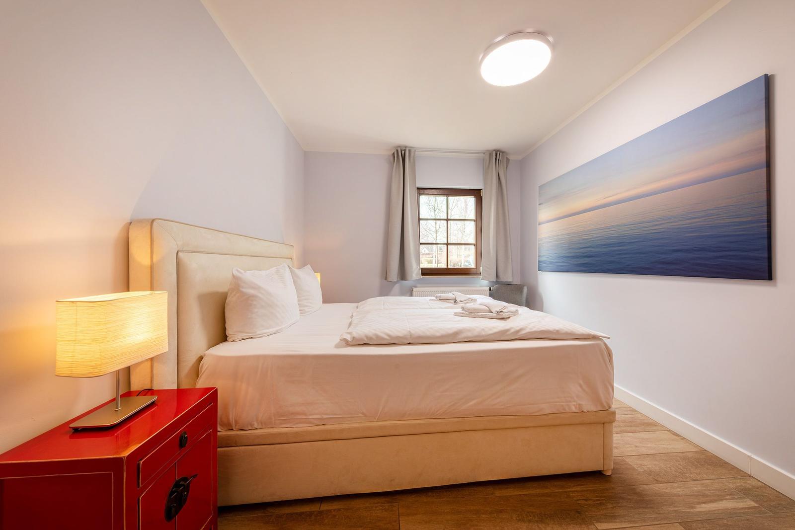 Schlafzimmer mit Bett, Nachttisch und Wandbild. Fenster mit Vorhängen.