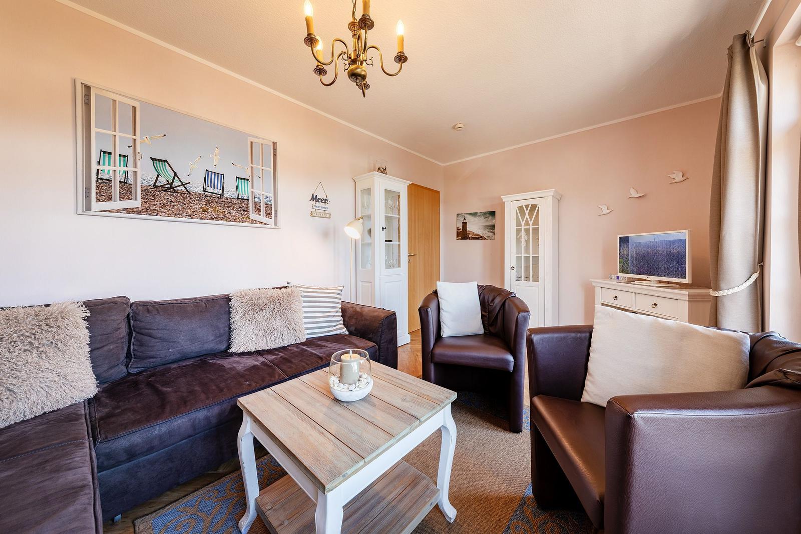 Wohnzimmer mit Sofa, Sesseln, Couchtisch und Fernseher. Wandbild zeigt Strand.
