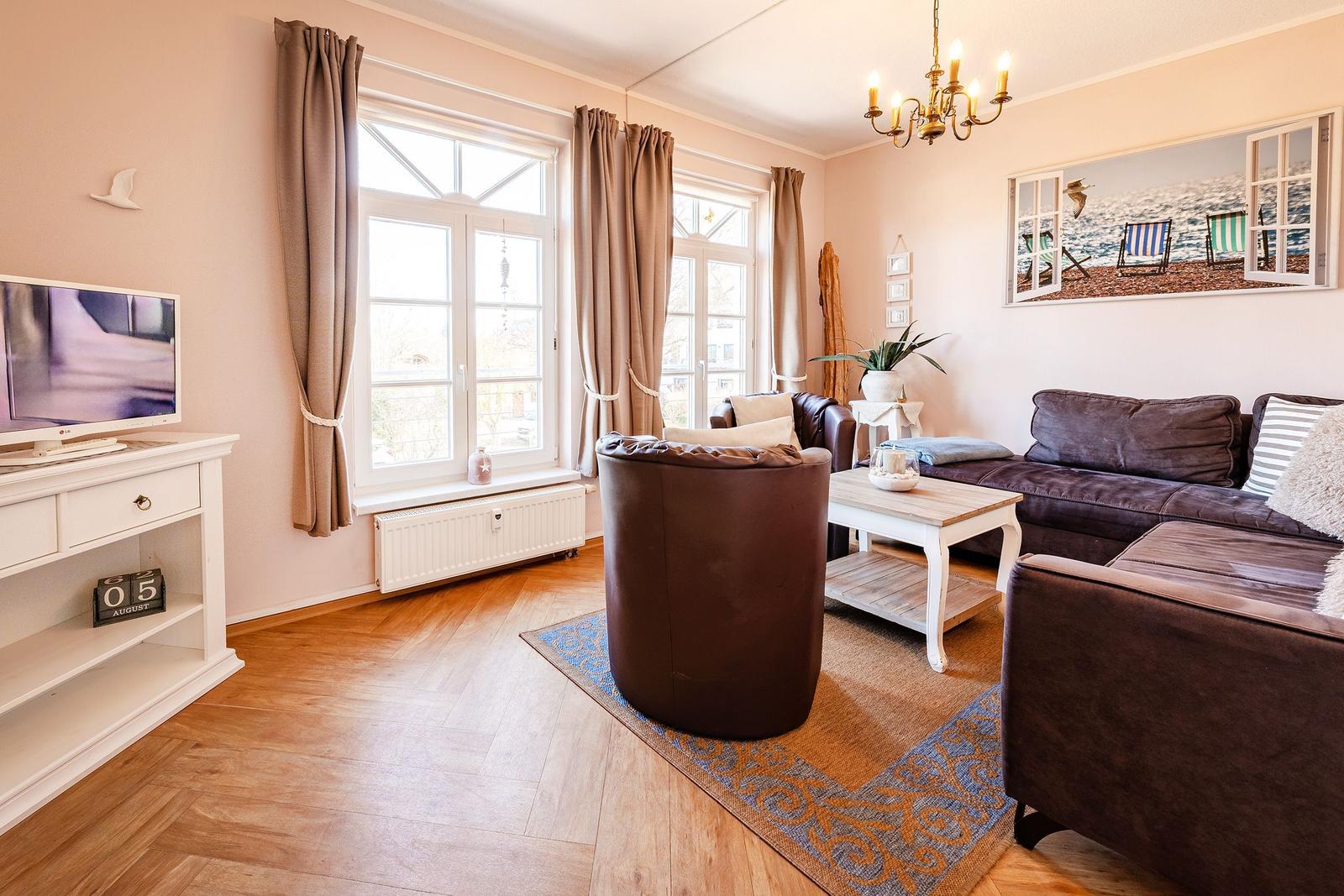 Wohnzimmer mit Sofa, Sessel, TV und Fenstern mit Vorhängen.