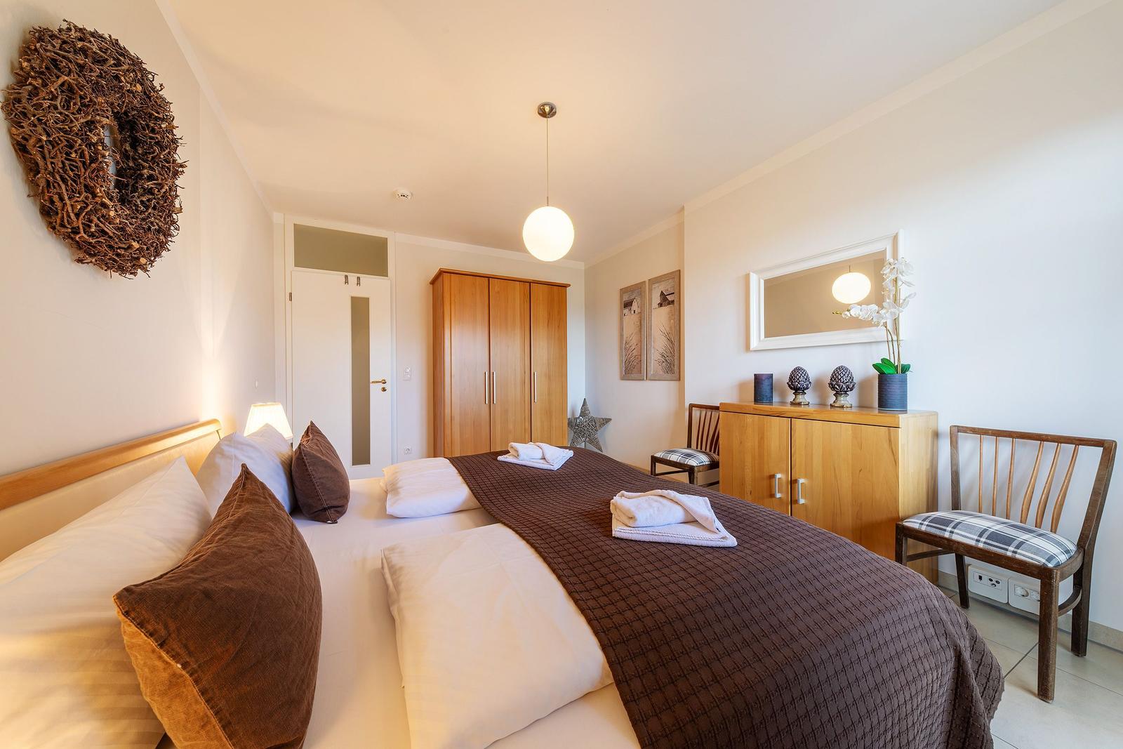 Schlafzimmer mit Bett, Kleiderschrank und Sitzgelegenheit. Wanddekoration und Spiegel sichtbar.