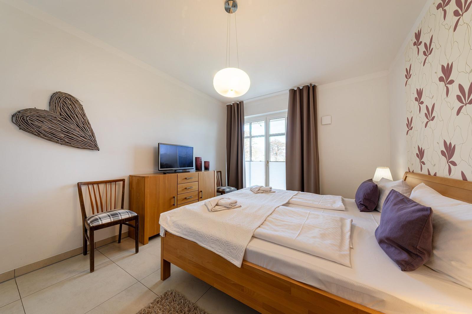 Schlafzimmer mit Bett, TV und Fenster. Wanddekoration und Holzmöbel.