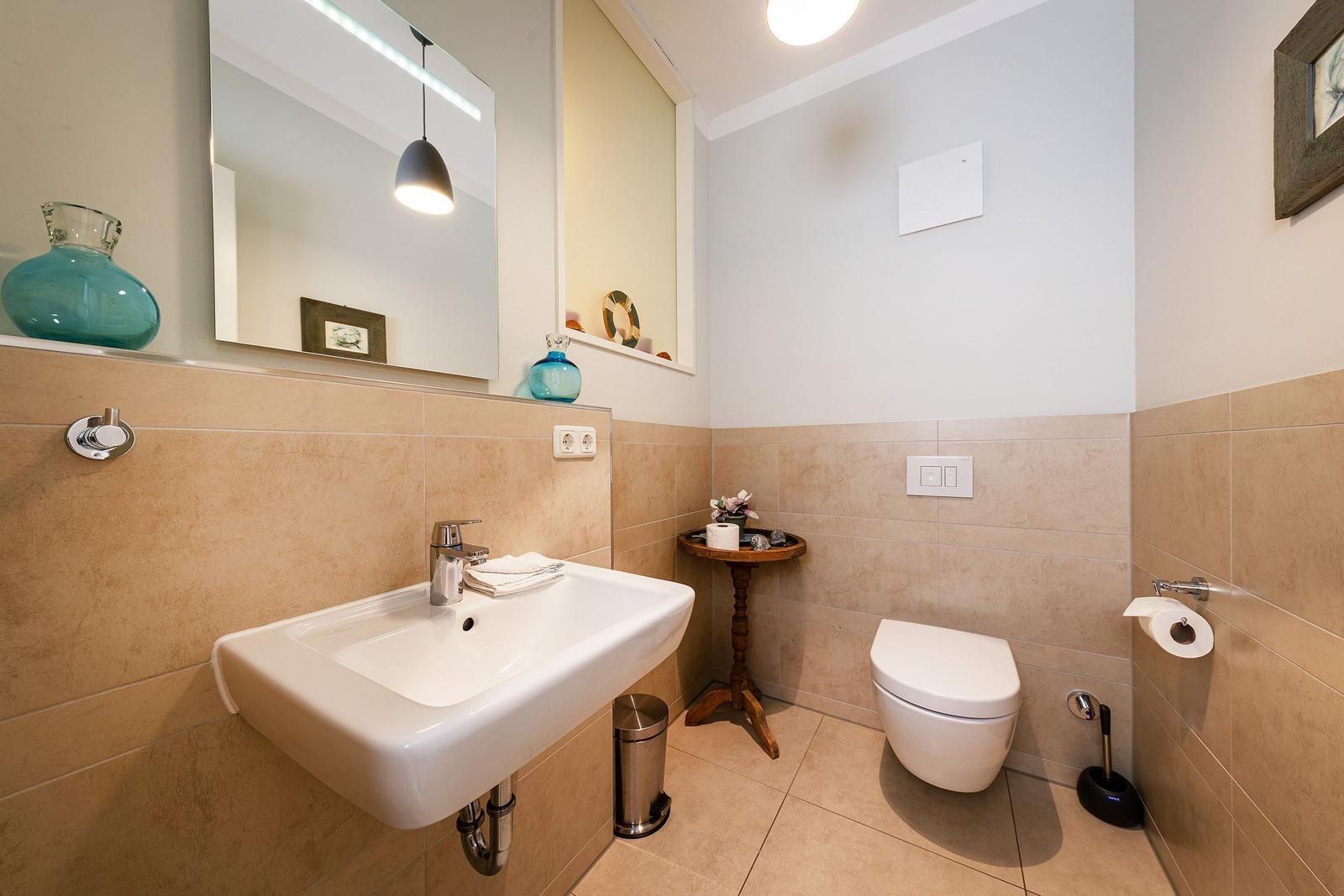 Badezimmer mit Waschbecken, Toilette und Spiegel. Wandverkleidung aus hellbraunen Fliesen.