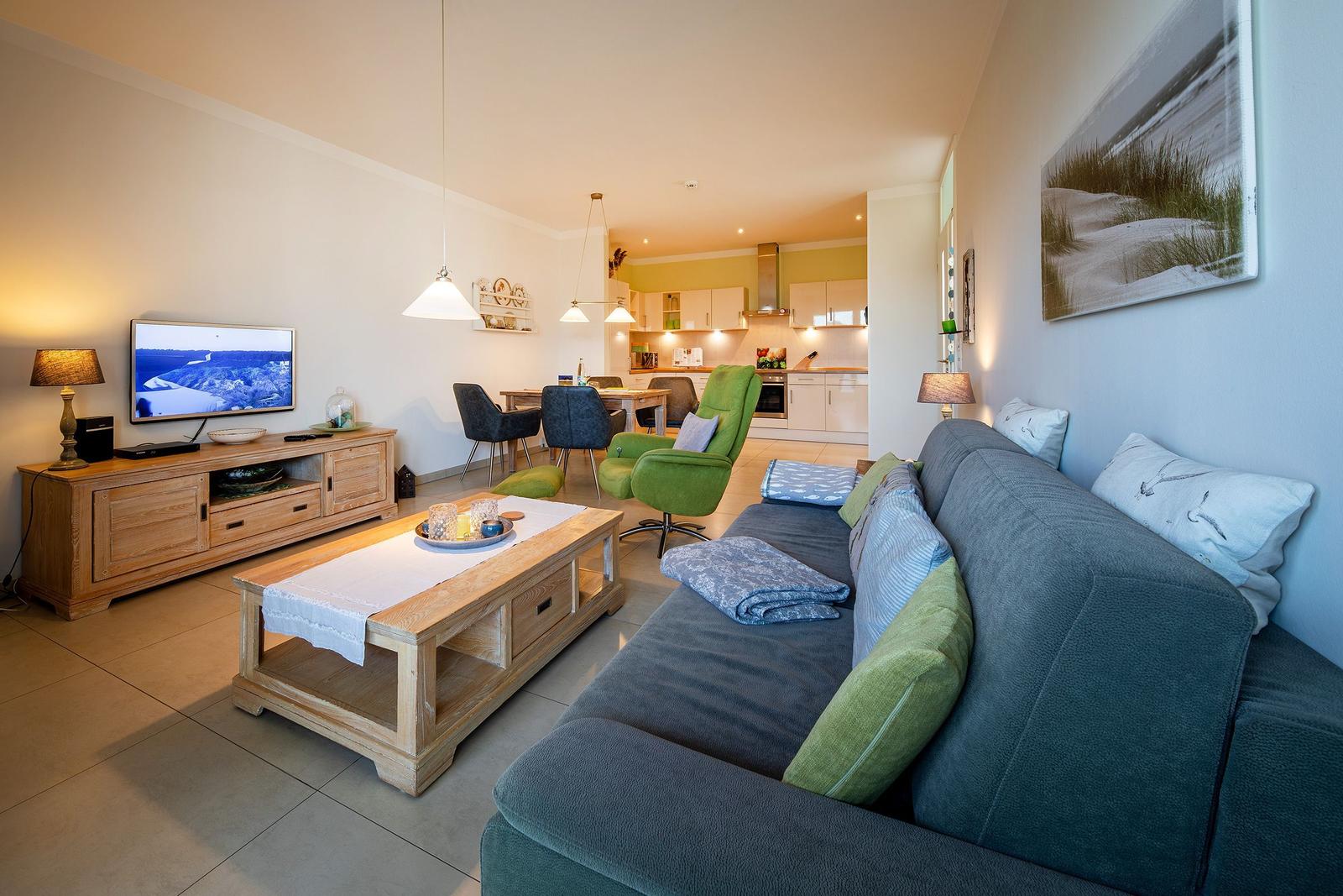 Wohnzimmer mit Sofa, Couchtisch und Fernseher. Küche und Essbereich im Hintergrund.
