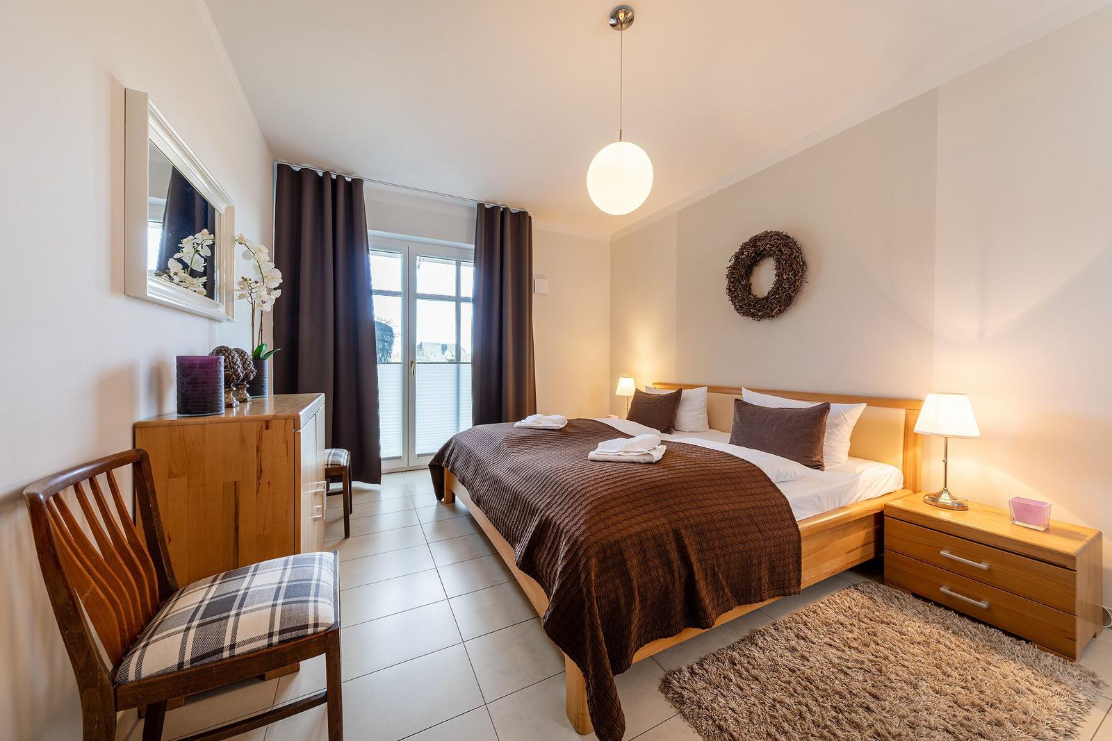 Schlafzimmer mit Bett, Nachttischen und Schreibtisch. Fenster mit Vorhängen und Deckenleuchte.