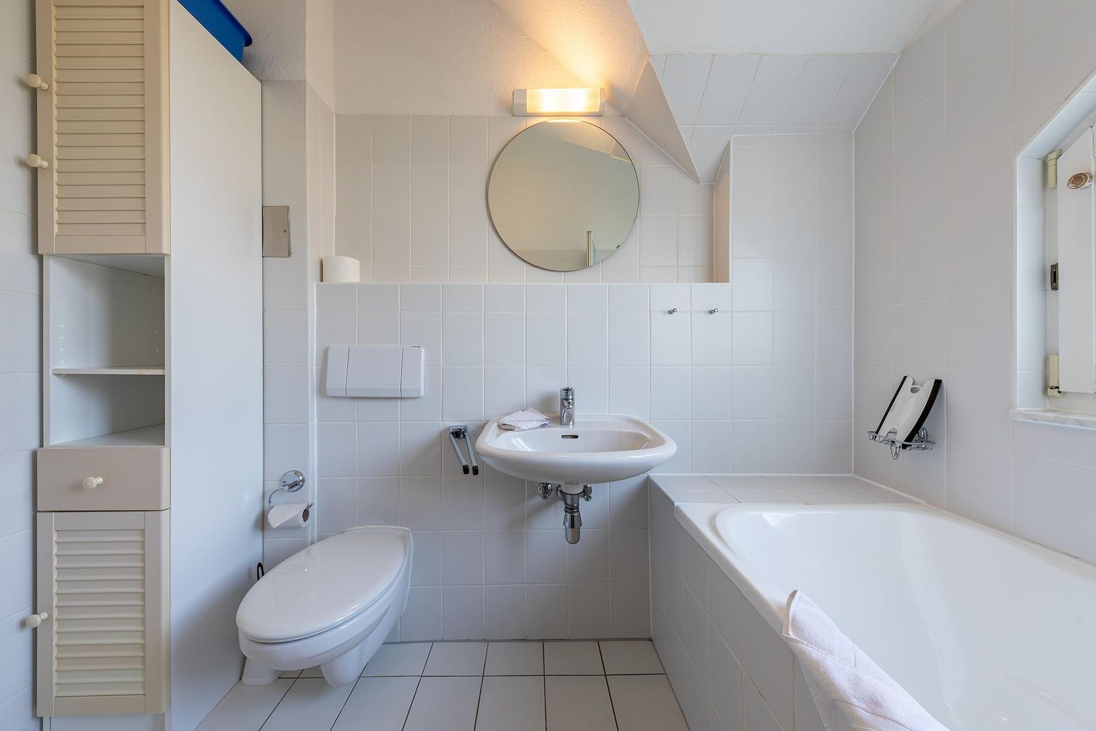 Badezimmer mit Badewanne, Waschbecken und Toilette. Weiße Fliesen und Spiegelschrank.