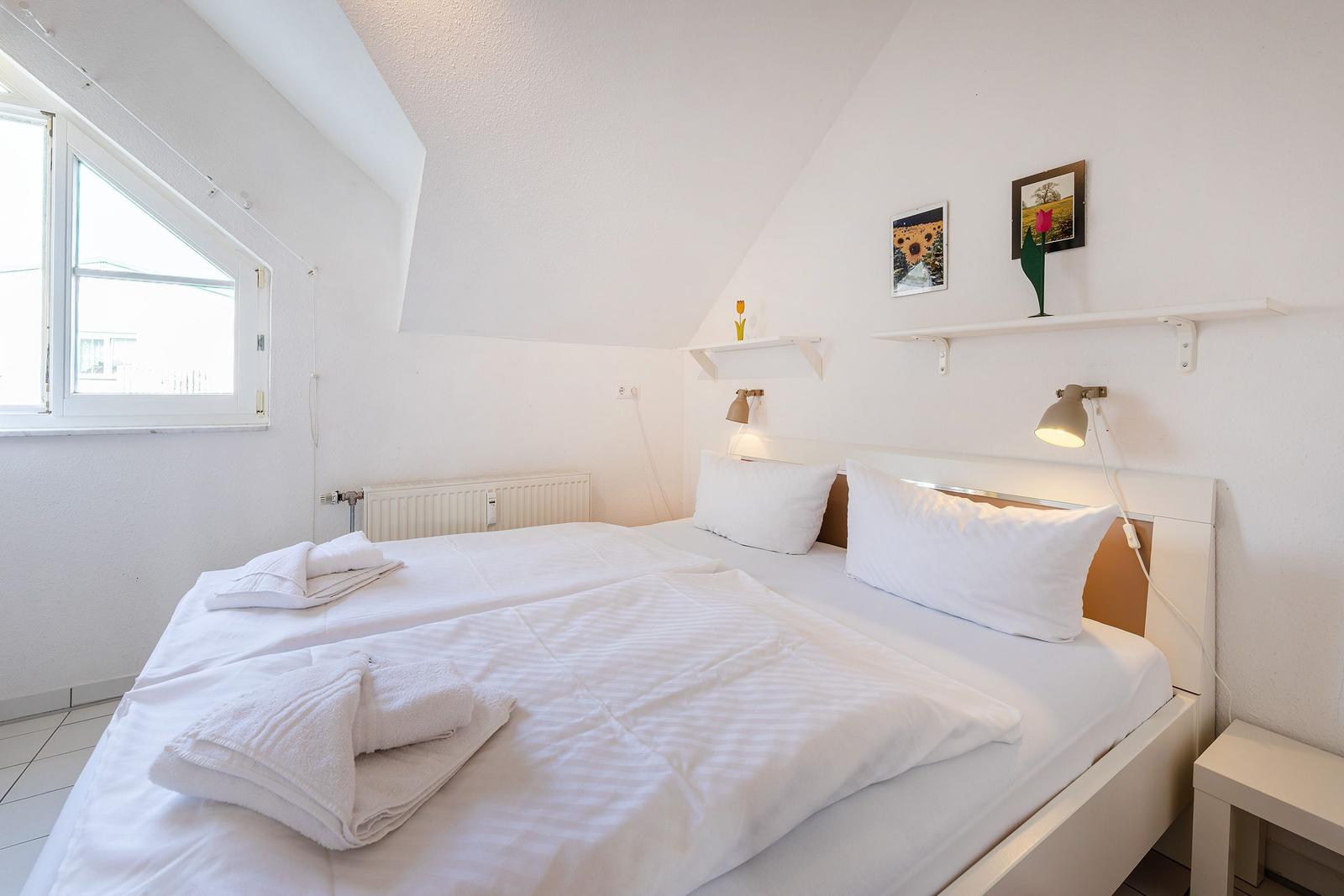 Schlafzimmer mit Doppelbett, Fenster und Wandregalen mit Bildern.