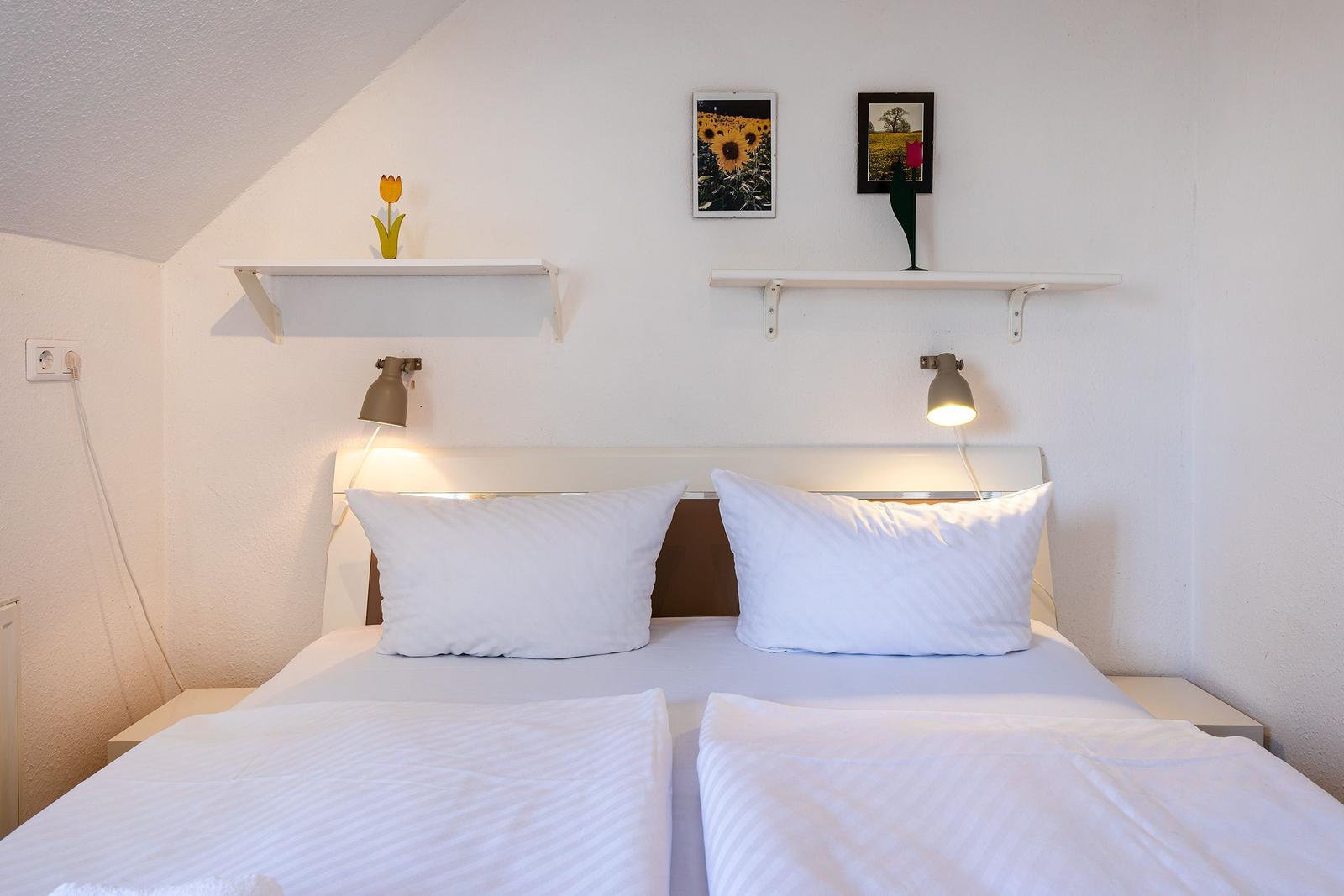 Ein Bett mit weißen Laken und Kissen. An der Wand hängen zwei Regale mit Bildern und Dekoration.