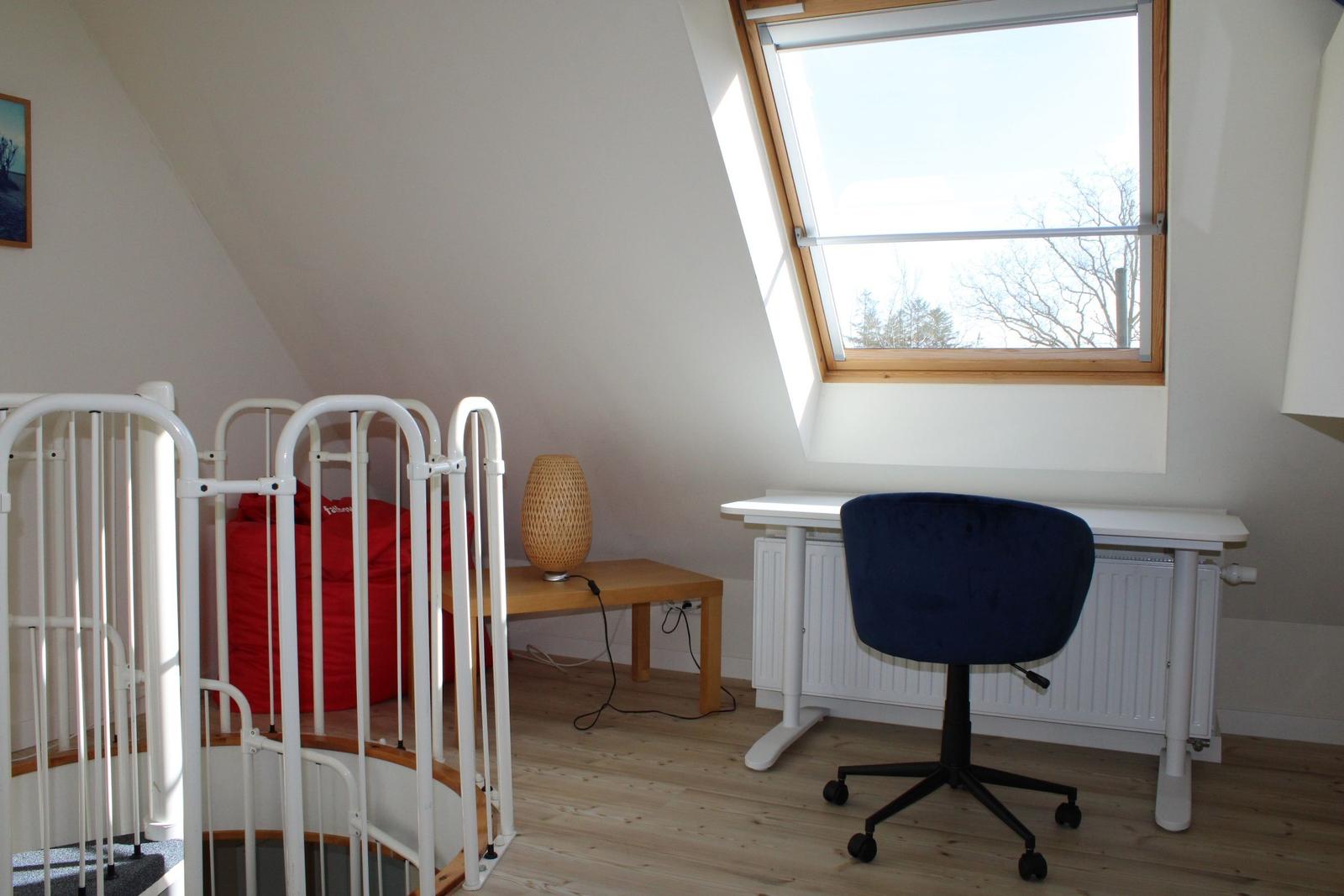 Schlafzimmer mit Schreibtisch, Stuhl und Fenster. Rote Matratze hinter Gitter.