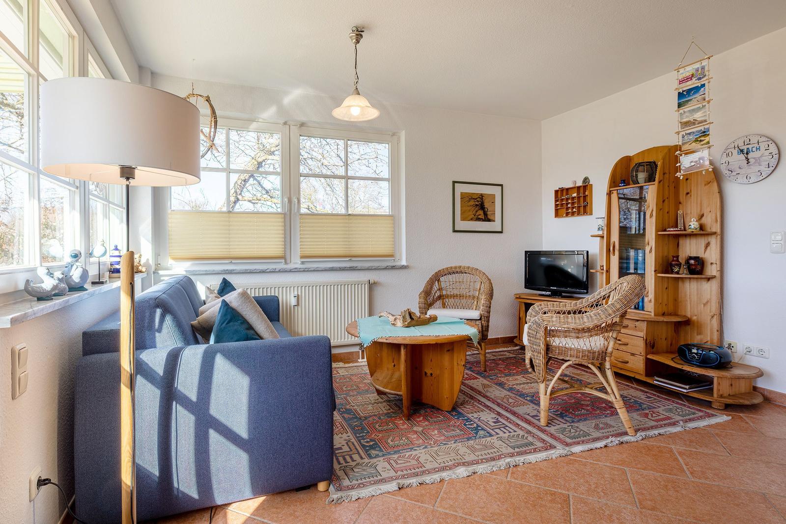 Wohnzimmer mit Sofa, zwei Rattanstühlen, Holztisch und Teppich. Fenster mit Jalousien und Holzregal.