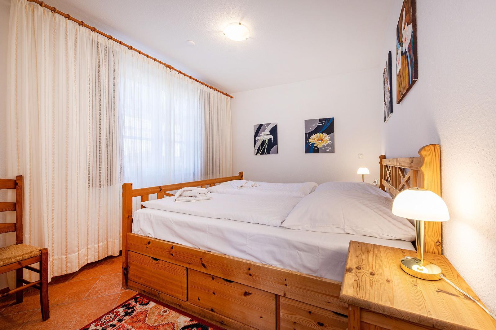 Schlafzimmer mit Holzbett, weißen Laken und Vorhängen. Nachttisch mit Lampe und Wandbildern.