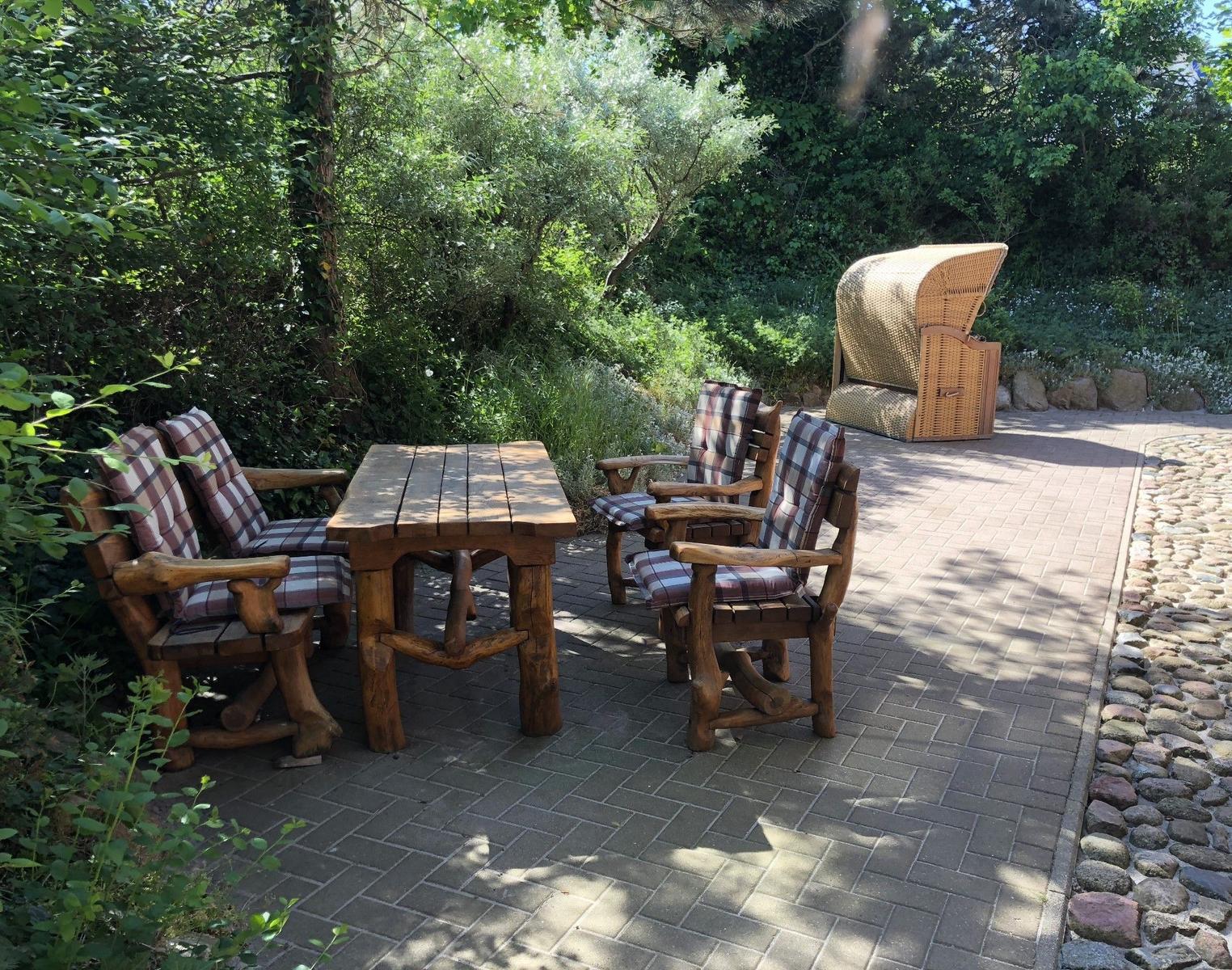 Terrasse mit Holztisch und vier Stühlen, daneben Strandkorb und grüne Umgebung.