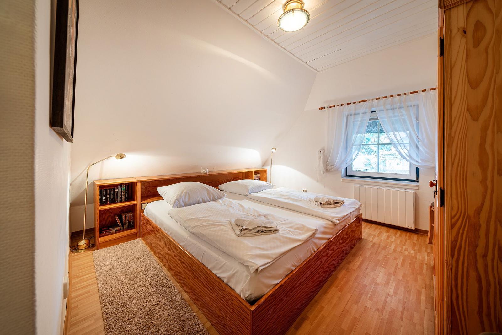Schlafzimmer mit Doppelbett, Holzboden und Fenster mit weißen Vorhängen.
