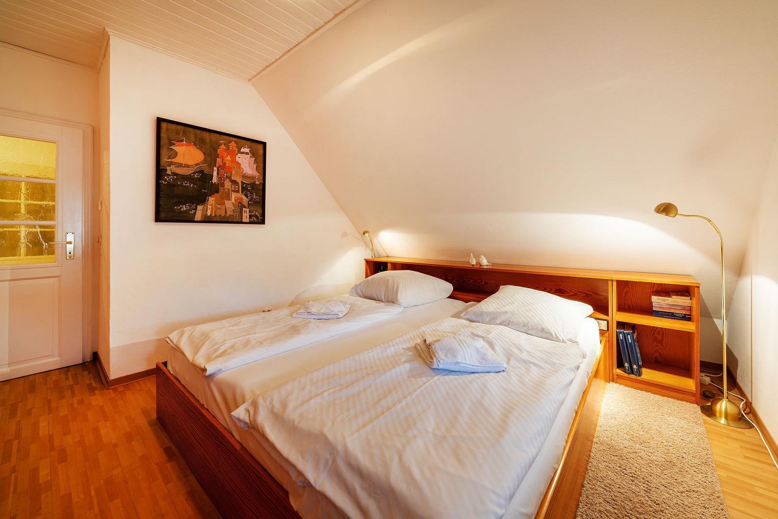 Schlafzimmer mit Doppelbett, Holzregal und Wandbild unter schräger Decke.